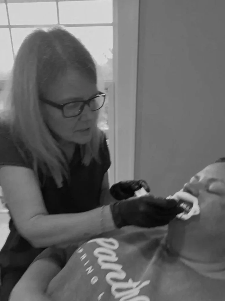 A woman wearing glasses and gloves appears to be examining or treating a person's mouth or face, who is lying down with their head tilted back, in an indoor setting with a window in the background.