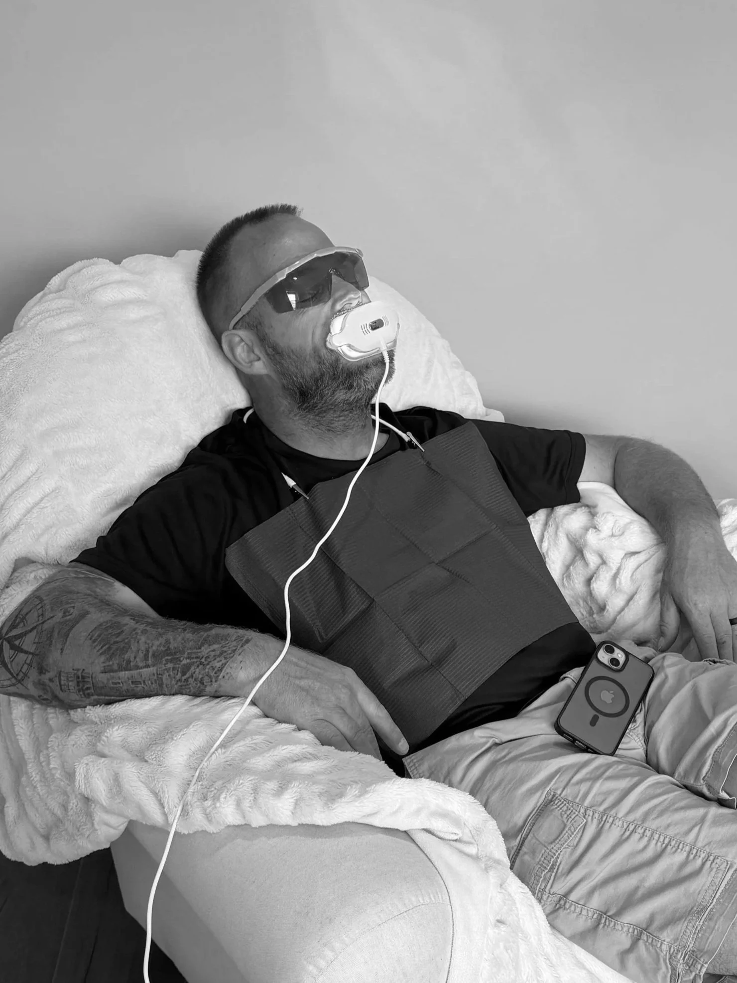 A man with a beard and tattoos on his left arm is lying in a hospital bed, wearing sunglasses and a sleep apnea mask connected to a ventilator. He has an iPhone with a case on his lap.