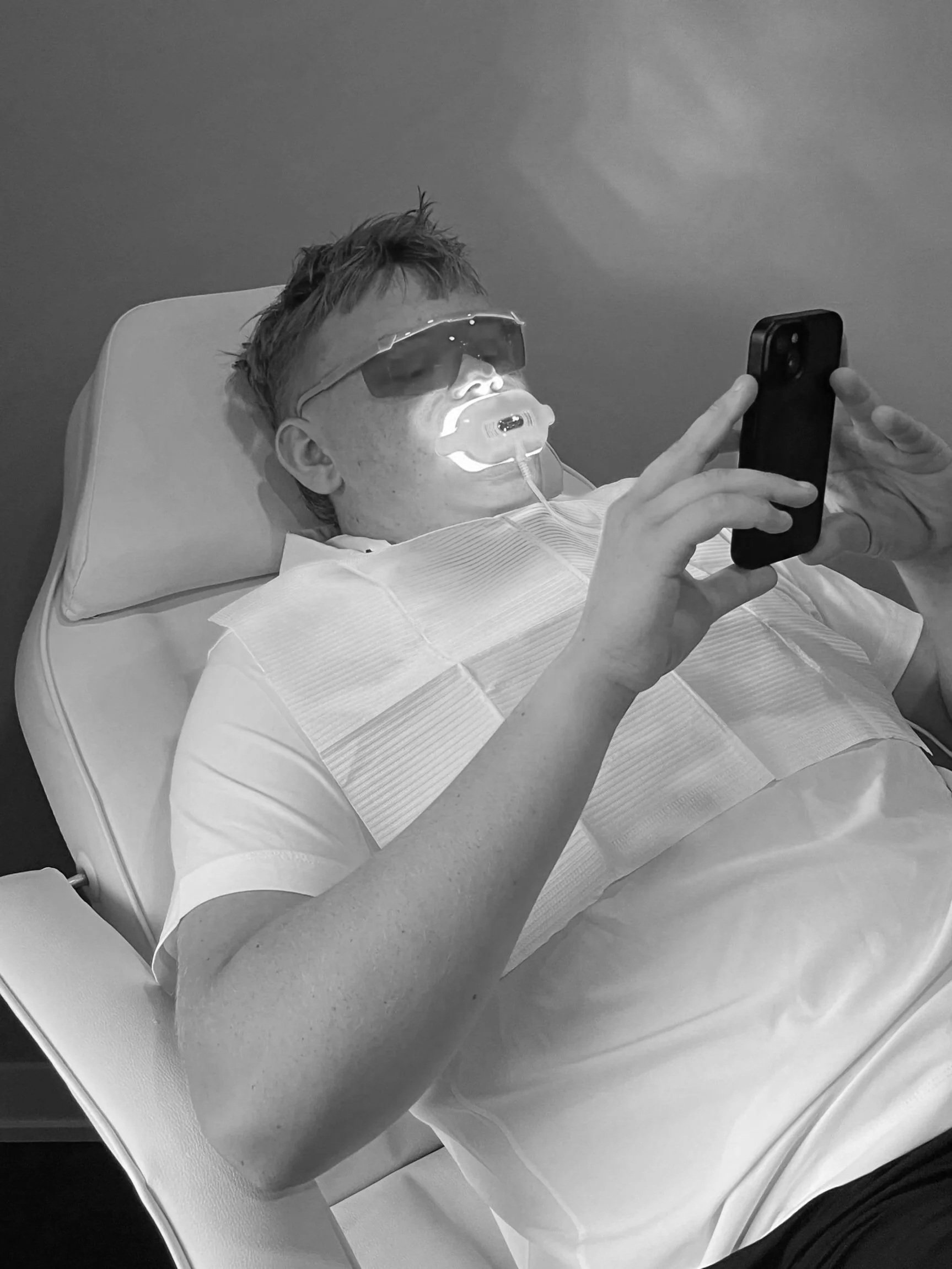 Person lying in a hospital bed with a medical mask and sunglasses, holding a smartphone.
