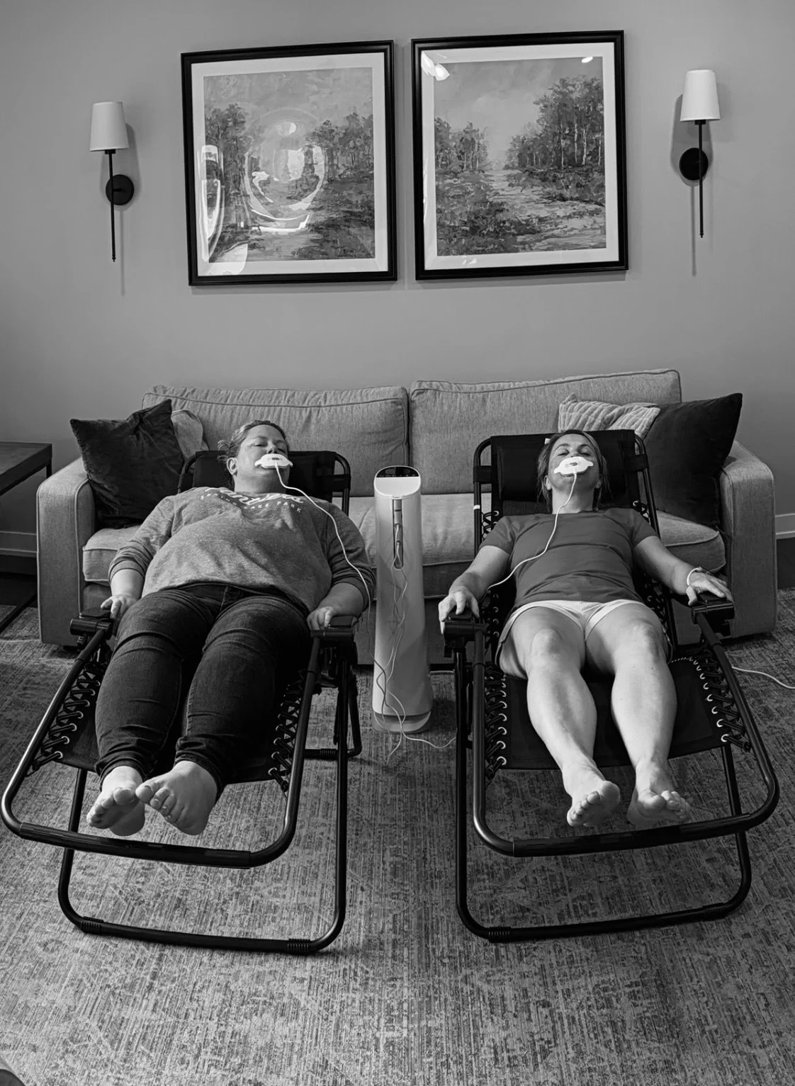 Two women lying on lounge chairs with medical devices on their faces, connected to a central machine, in a living room with wall-mounted lamps and framed landscape paintings.