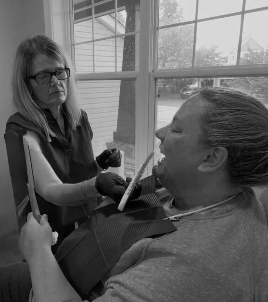 Woman wearing glasses and gloves checks a woman's teeth using a dental mirror while she reclines in a dental chair, near a window.