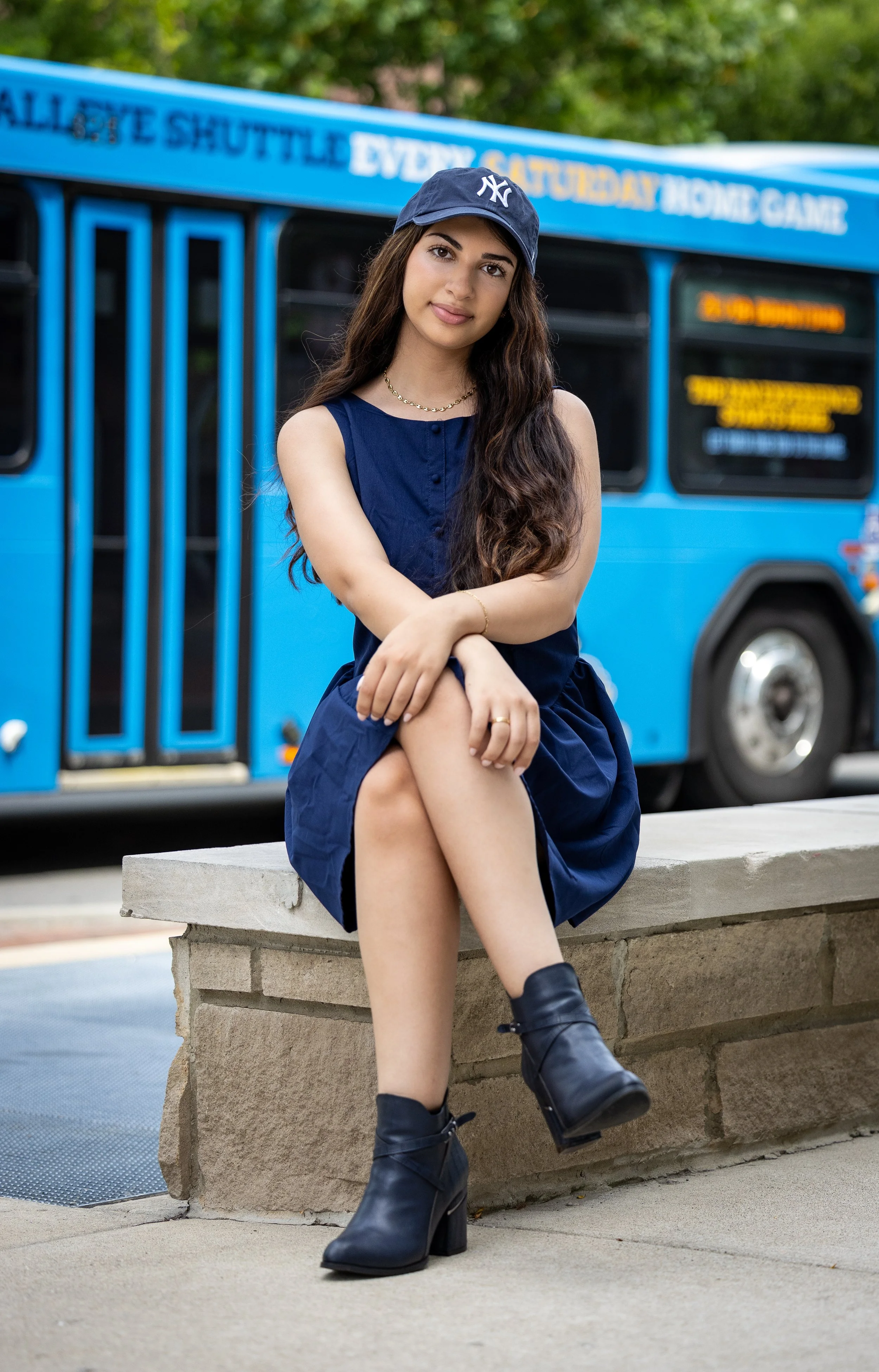 A young woman with long brown hair, wearing a navy blue dress, black heeled boots, and a navy cap with a NYC logo, sitting on a stone bench in front of a blue bus.