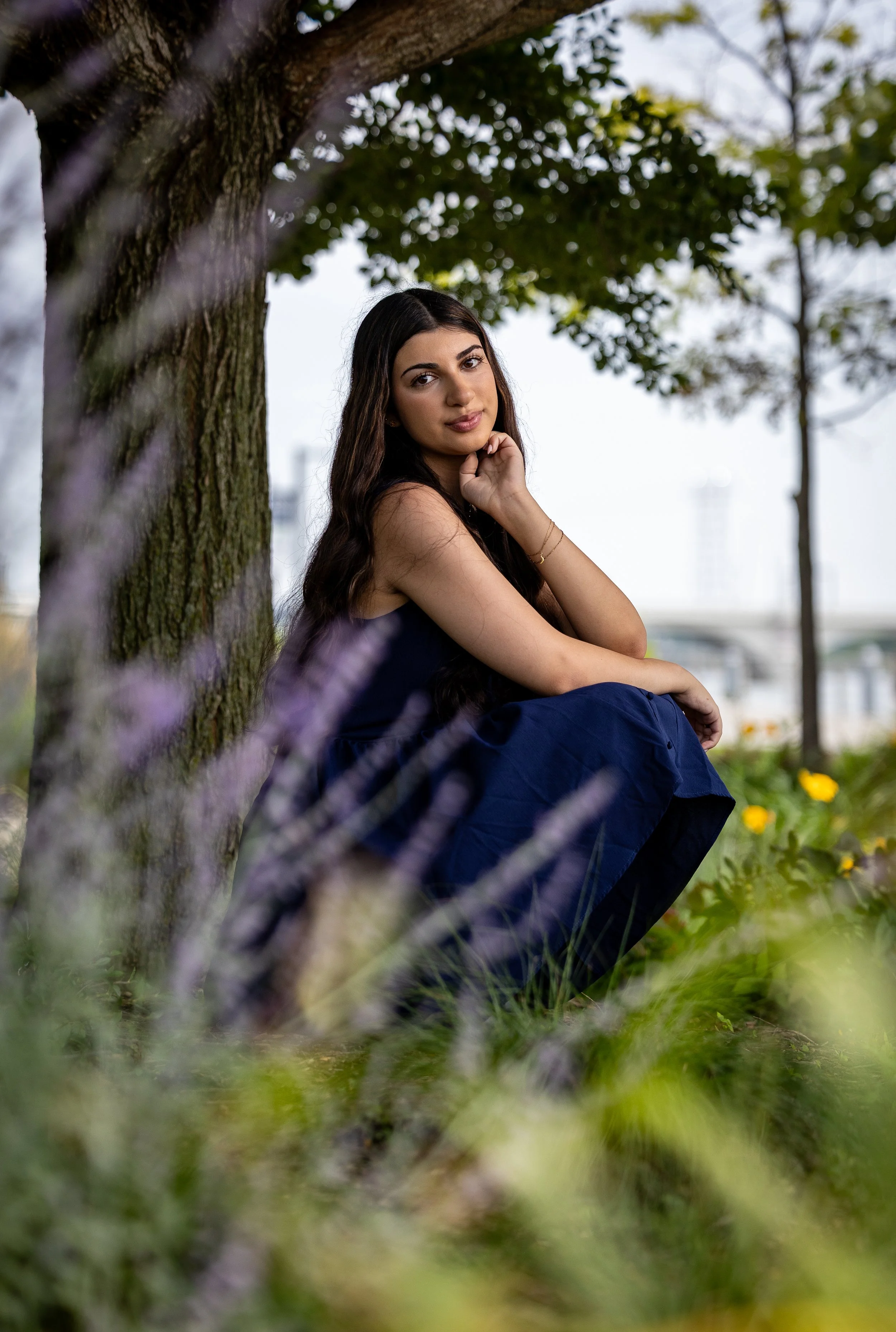 A woman sitting outdoors under a tree, smiling gently, with blurred foreground of purple flowers and greenery.