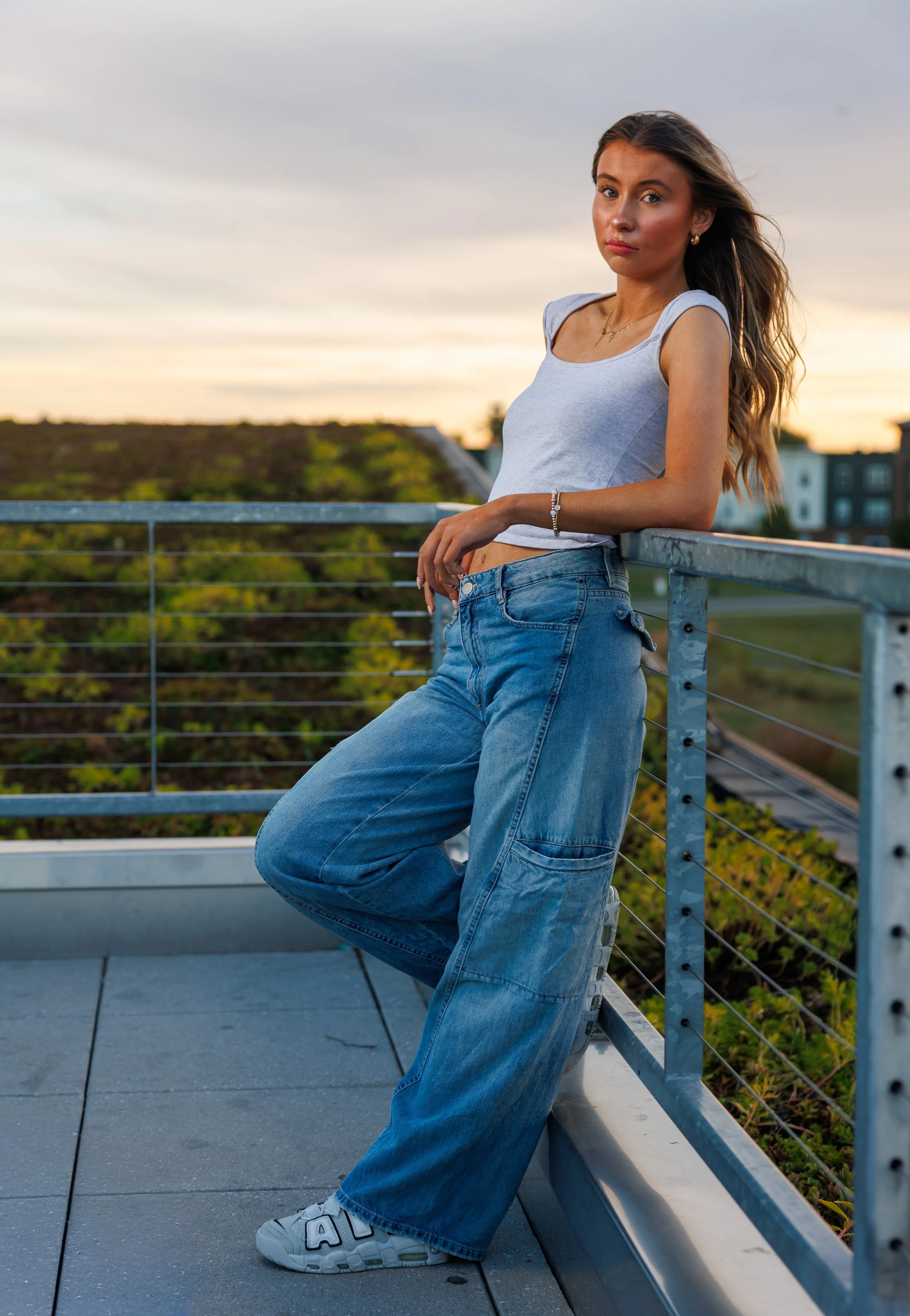 Young woman standing on a rooftop balcony during sunset, leaning against a metal railing with her right hand, wearing a gray crop top, loose blue jeans, white sneakers, and jewelry, with buildings and greenery in the background.