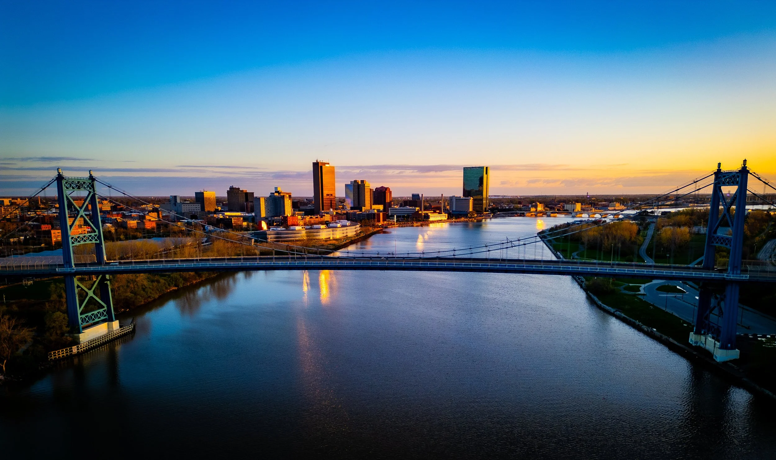 A city skyline at sunset with a river in the foreground and a bridge crossing over the river.