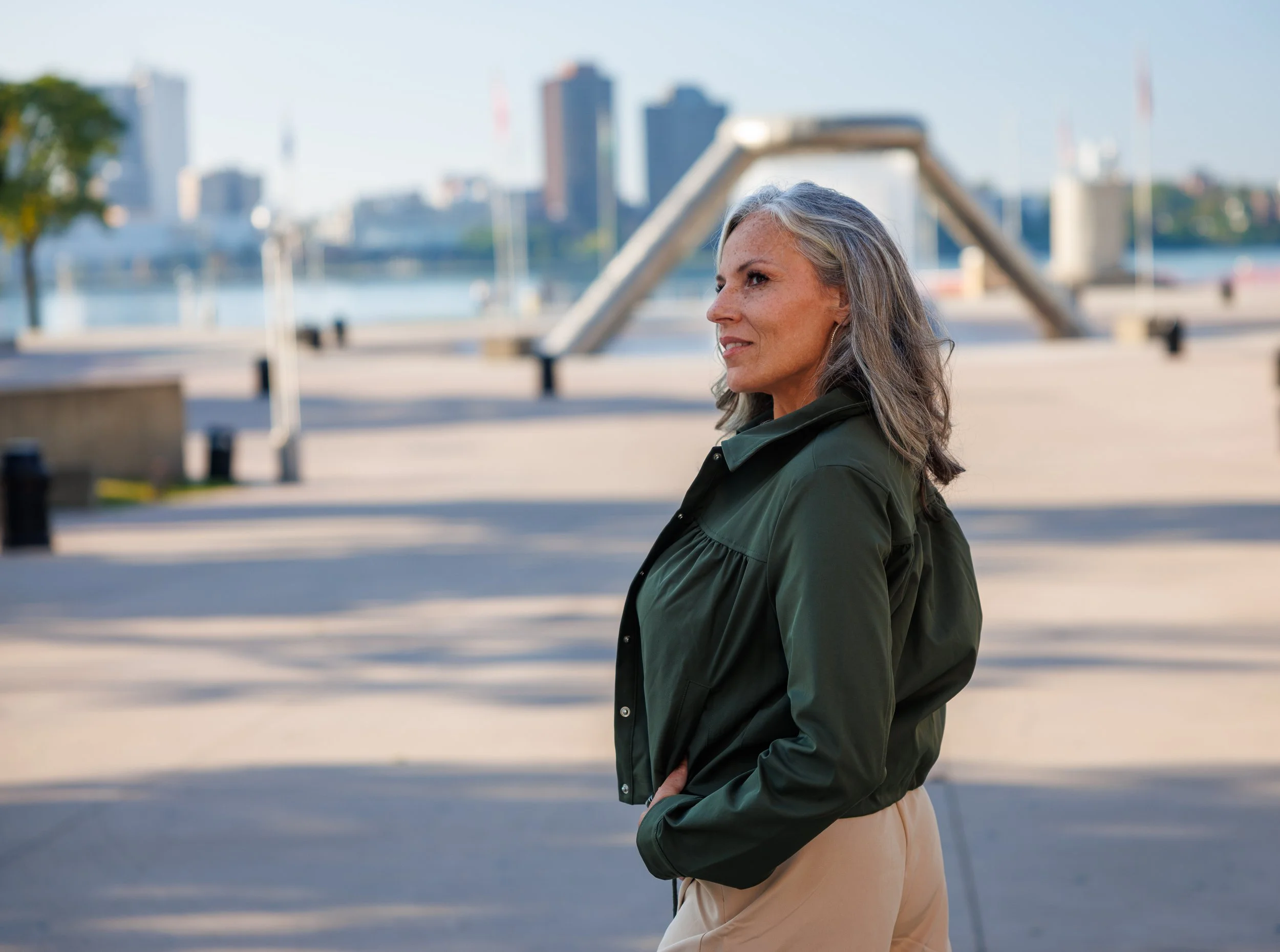 A woman with gray hair and wearing a green jacket standing outdoors near a waterfront with city buildings in the background.