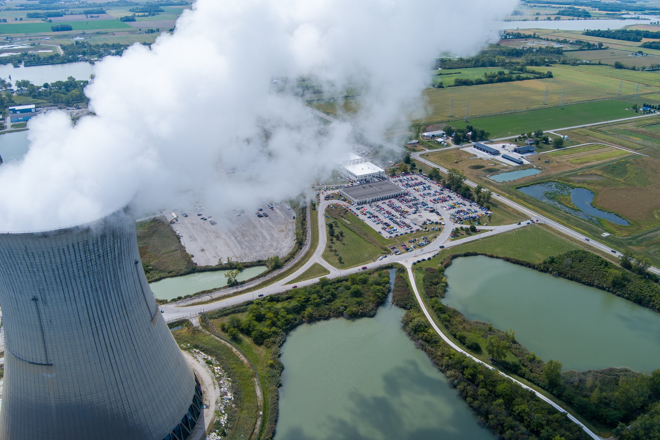 An aerial view of a nuclear power plant showing a large cooling tower emitting steam, surrounded by green fields, ponds, and some buildings and parking lots.