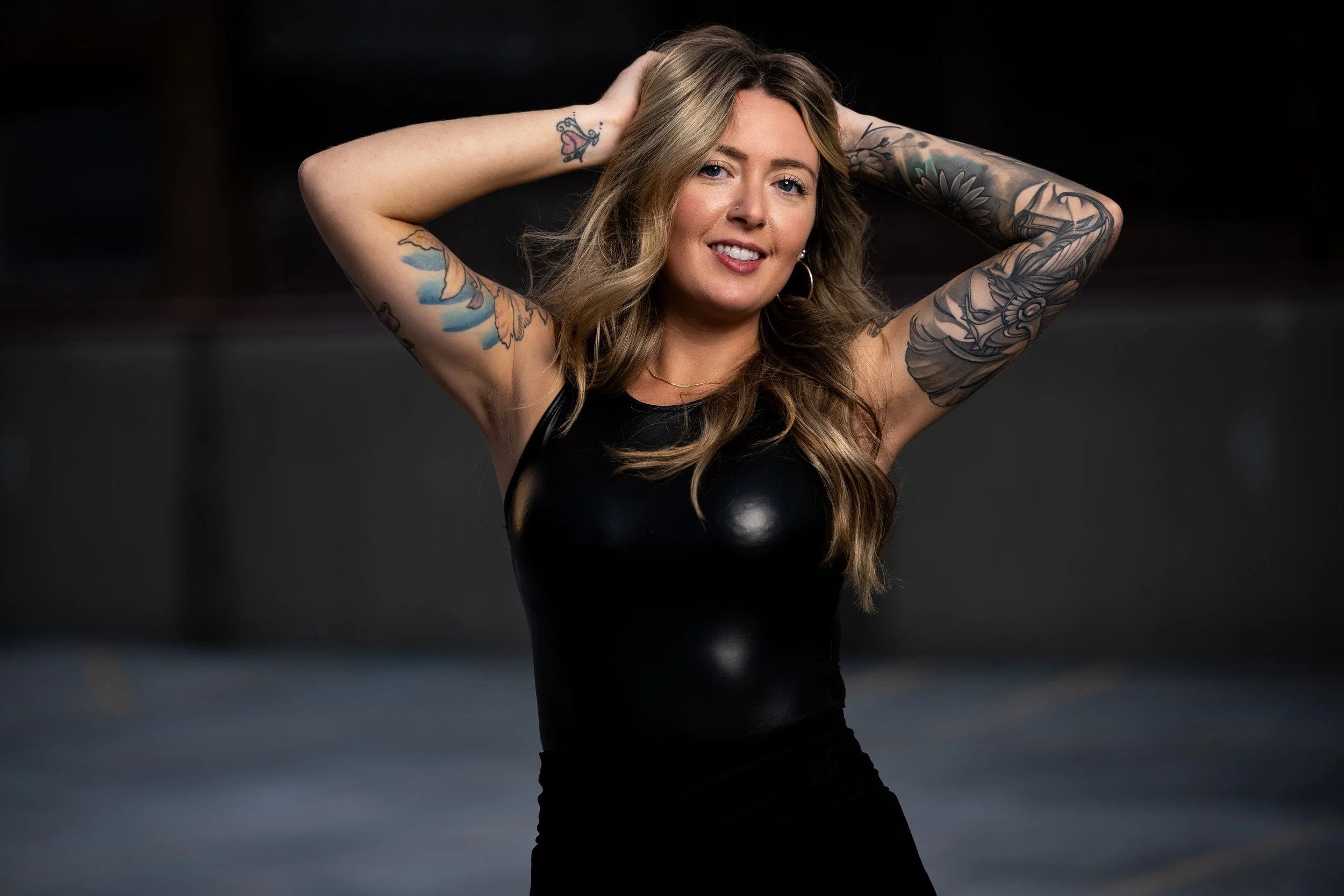 A woman with long wavy hair, tattoos on her arms, and hoop earrings smiling with her hands behind her head, wearing a black sleeveless top and standing against a dark background.