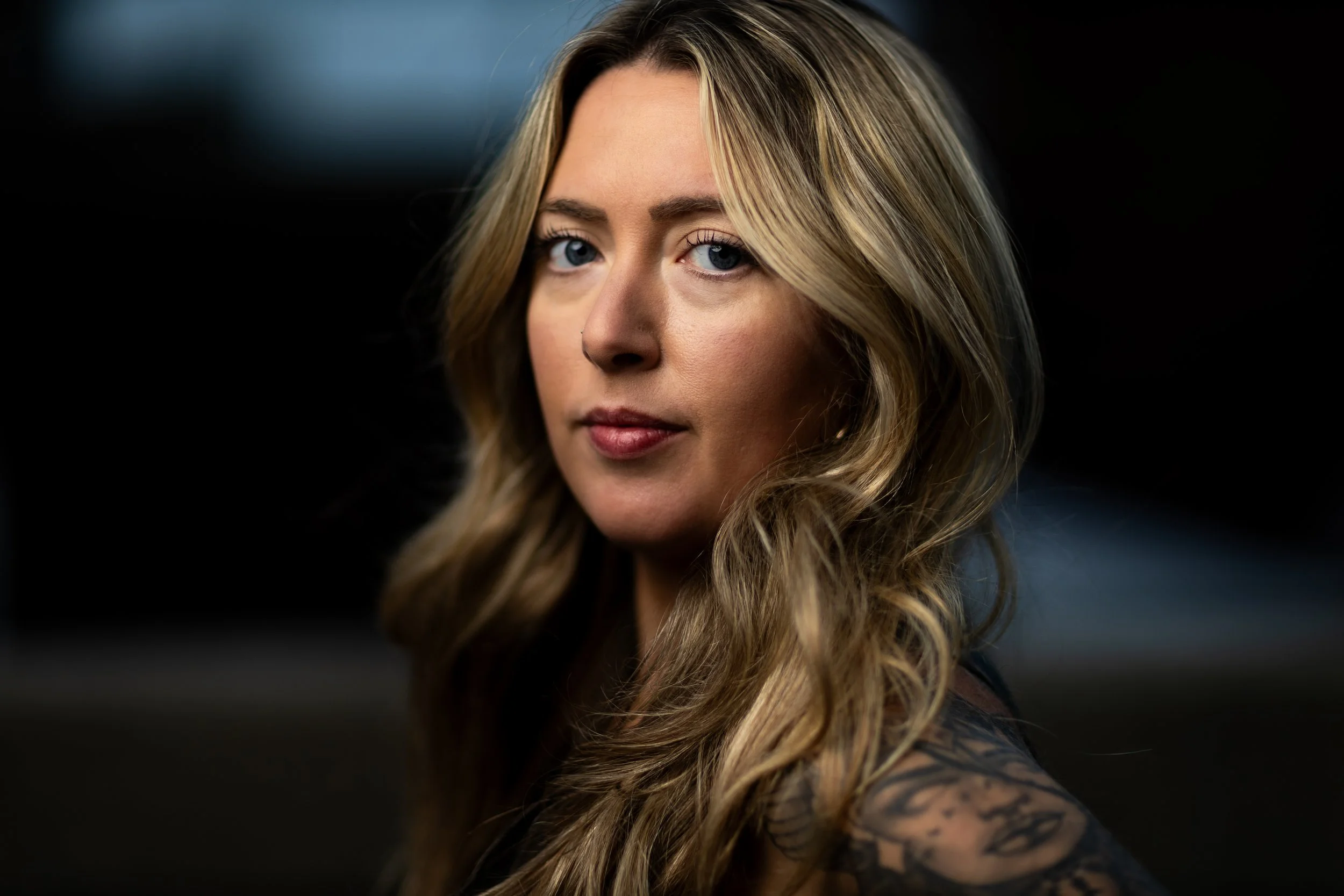 Close-up of a woman with long, wavy blonde hair and blue eyes, looking at the camera with a serious expression. She has natural makeup, and the background is dark and blurred.