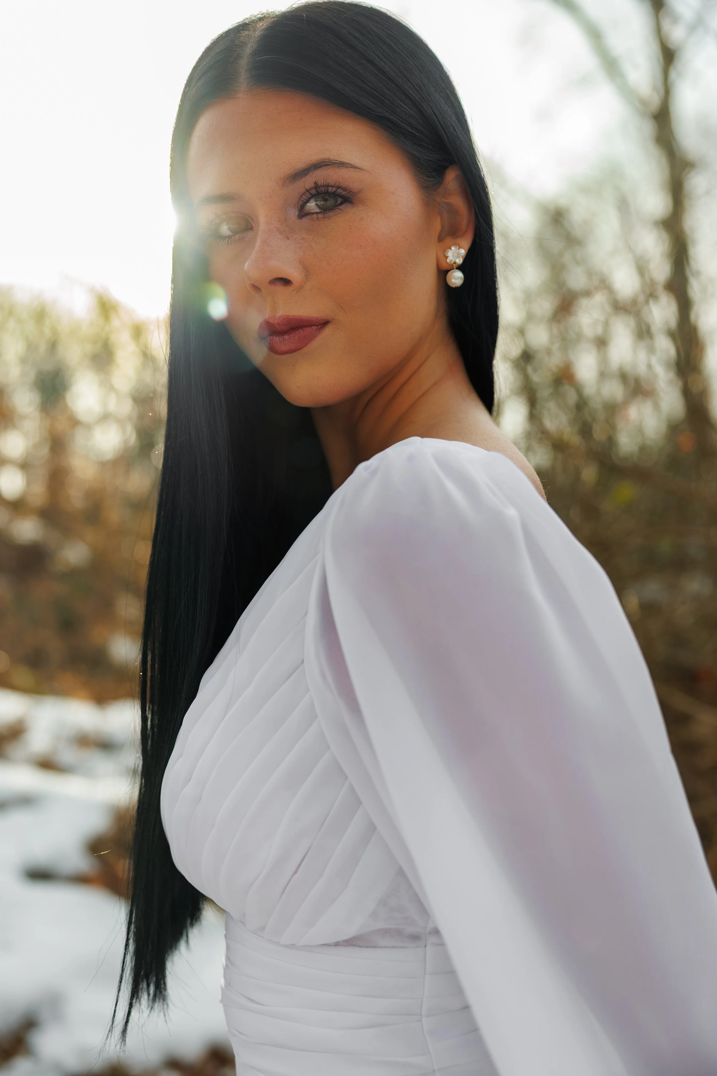 Close-up of a woman with long black hair, wearing pearl and floral earrings, in winter outdoor setting with snow and trees in the background.