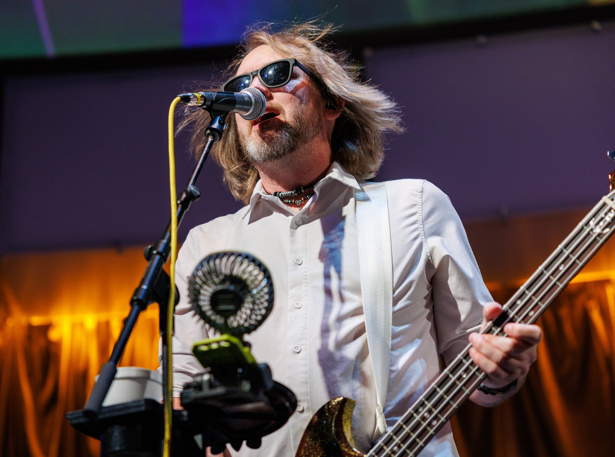 A man with long hair, sunglasses, and a beard singing into a microphone while playing an electric guitar on stage.