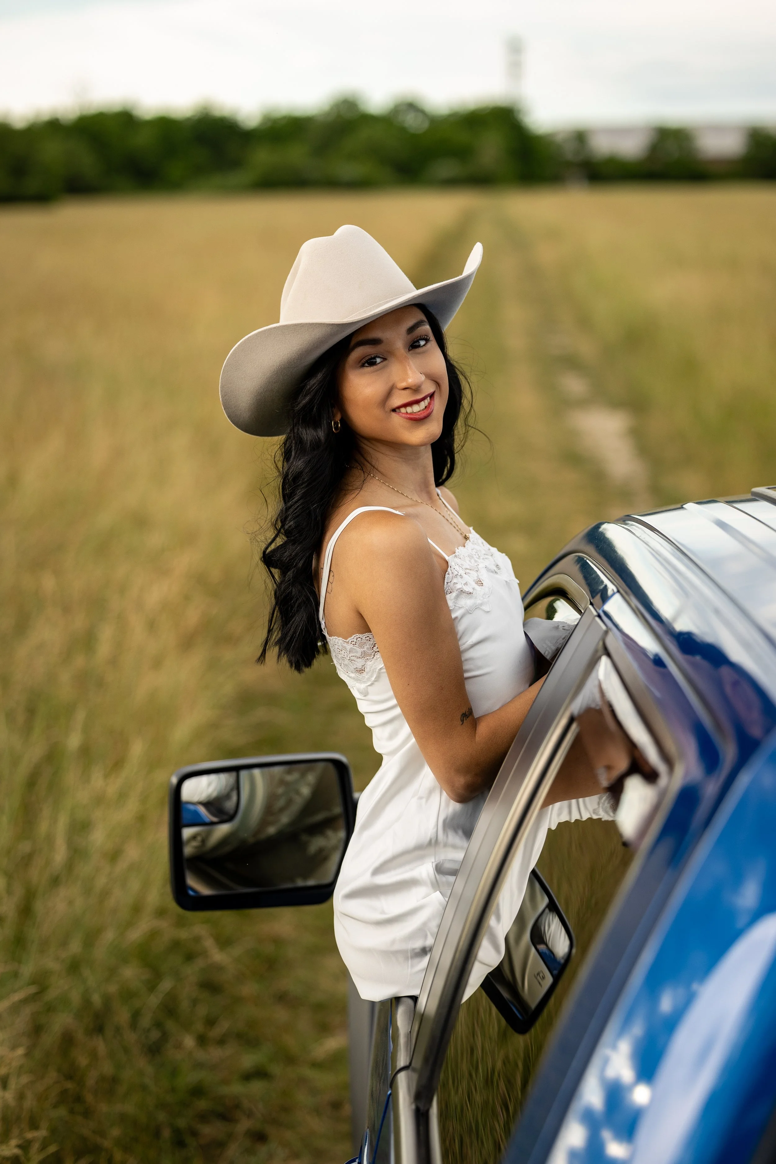 A woman with long dark hair, wearing a white dress and a large wide-brimmed white hat, standing partially inside a blue vehicle in a grassy field, smiling at the camera with a background of trees and a cloudy sky.