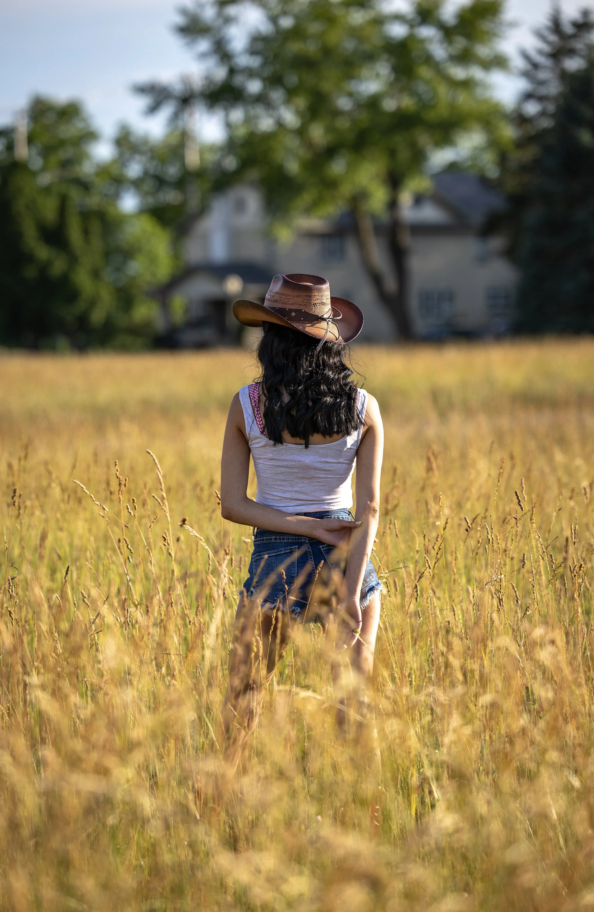 A woman in a tank top and denim shorts standing in a wheat field, wearing a cowboy hat, with trees and a house in the background on a sunny day.