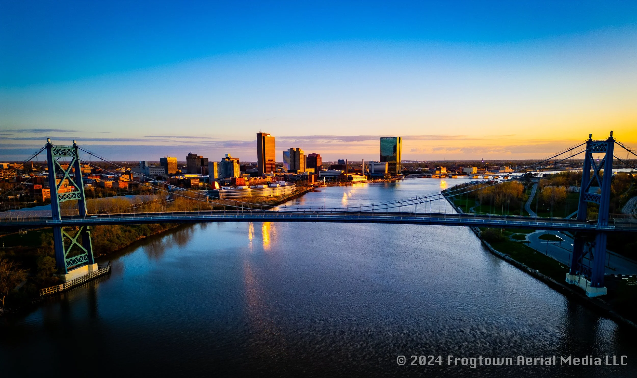 Aerial view of a city skyline with tall buildings, river, and a blue suspension bridge at sunset.