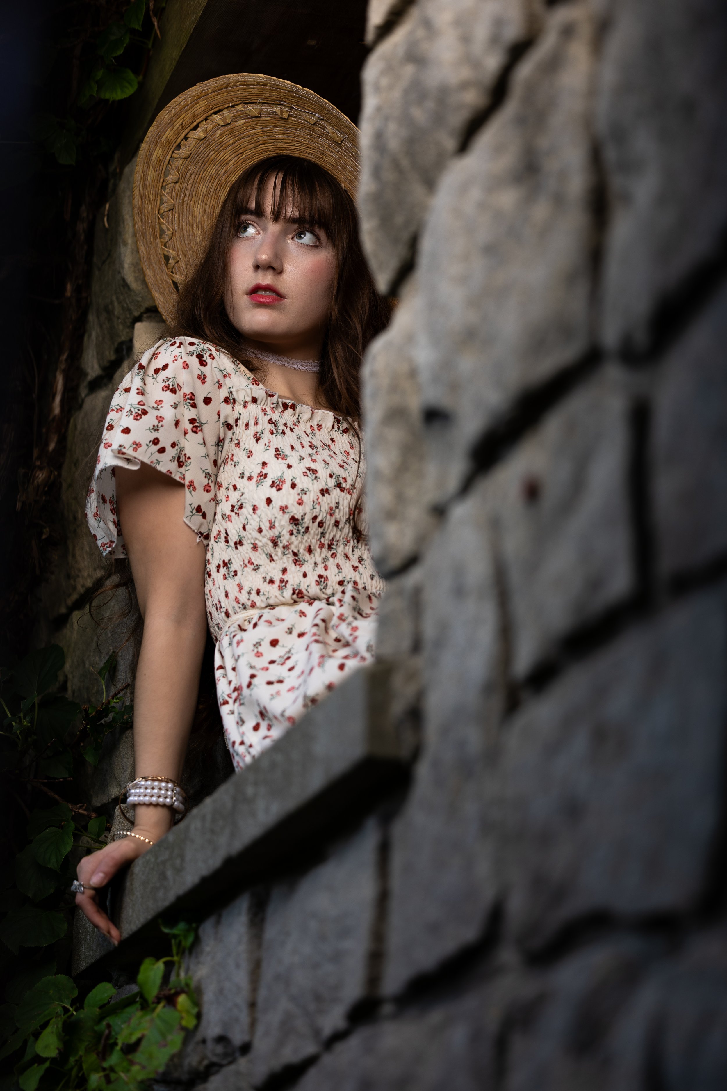 A young woman with brown hair and bangs wearing a wide-brimmed straw hat and a floral dress, leaning against a stone wall with greenery behind her, looking upward and to the side.