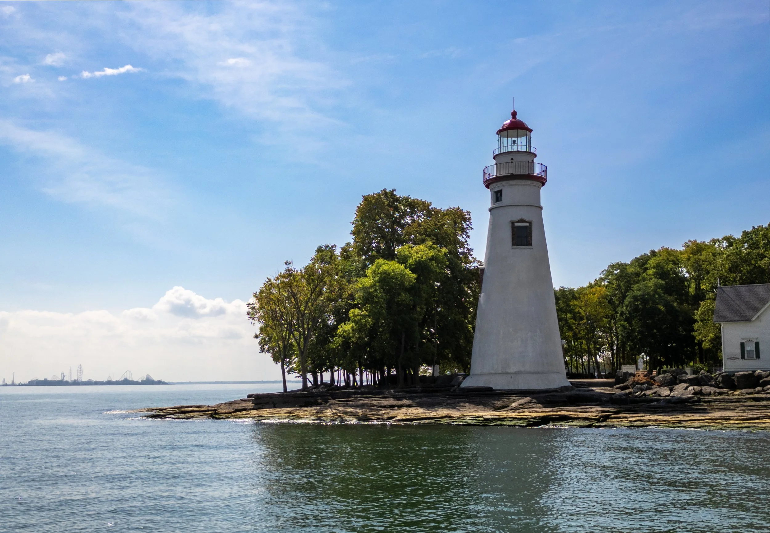 A lighthouse with a red top and white body stands next to a building on a rocky shoreline, surrounded by green trees, with the water and a distant city skyline in the background under a partly cloudy sky.