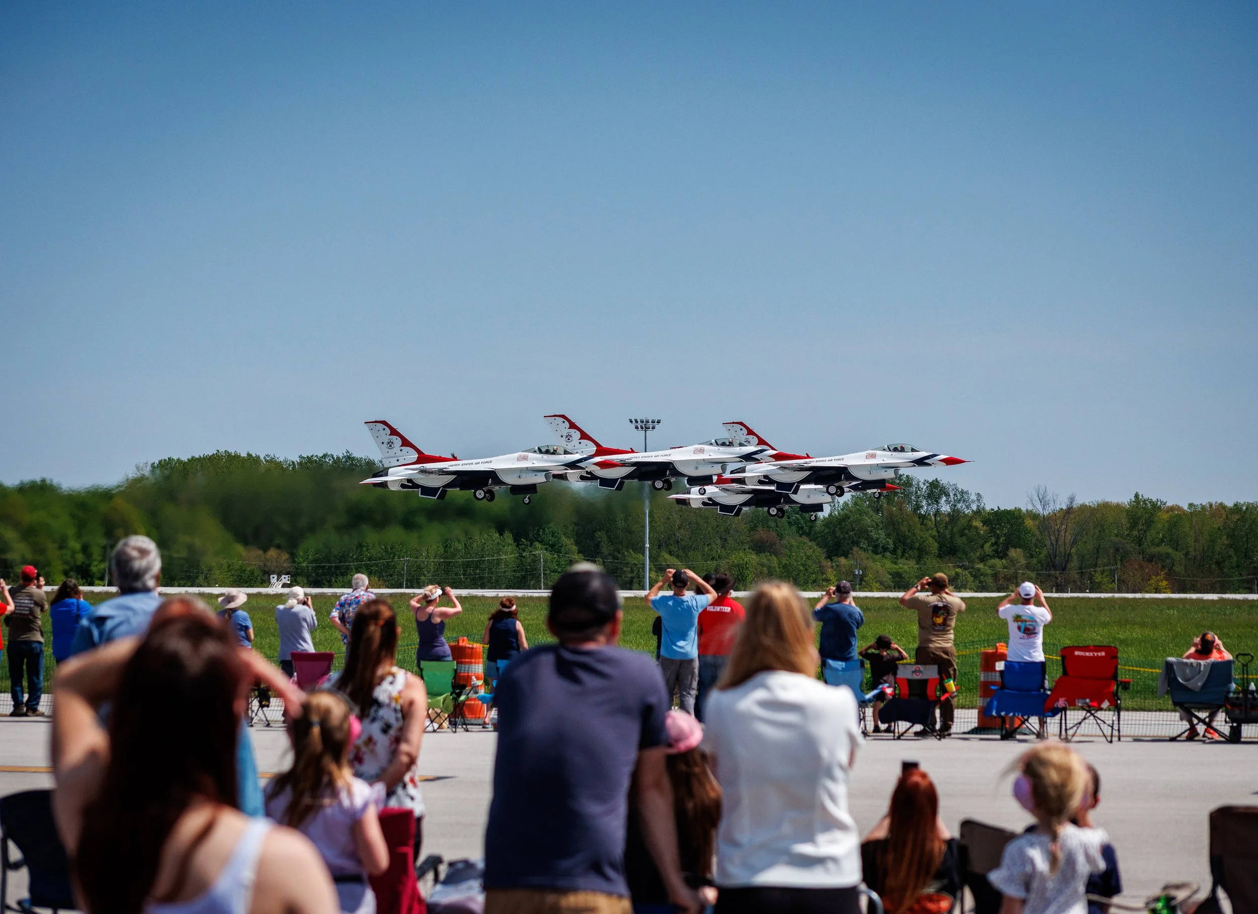 Several fighter jets fly in unison during an air show, with a crowd of spectators watching from the ground.
