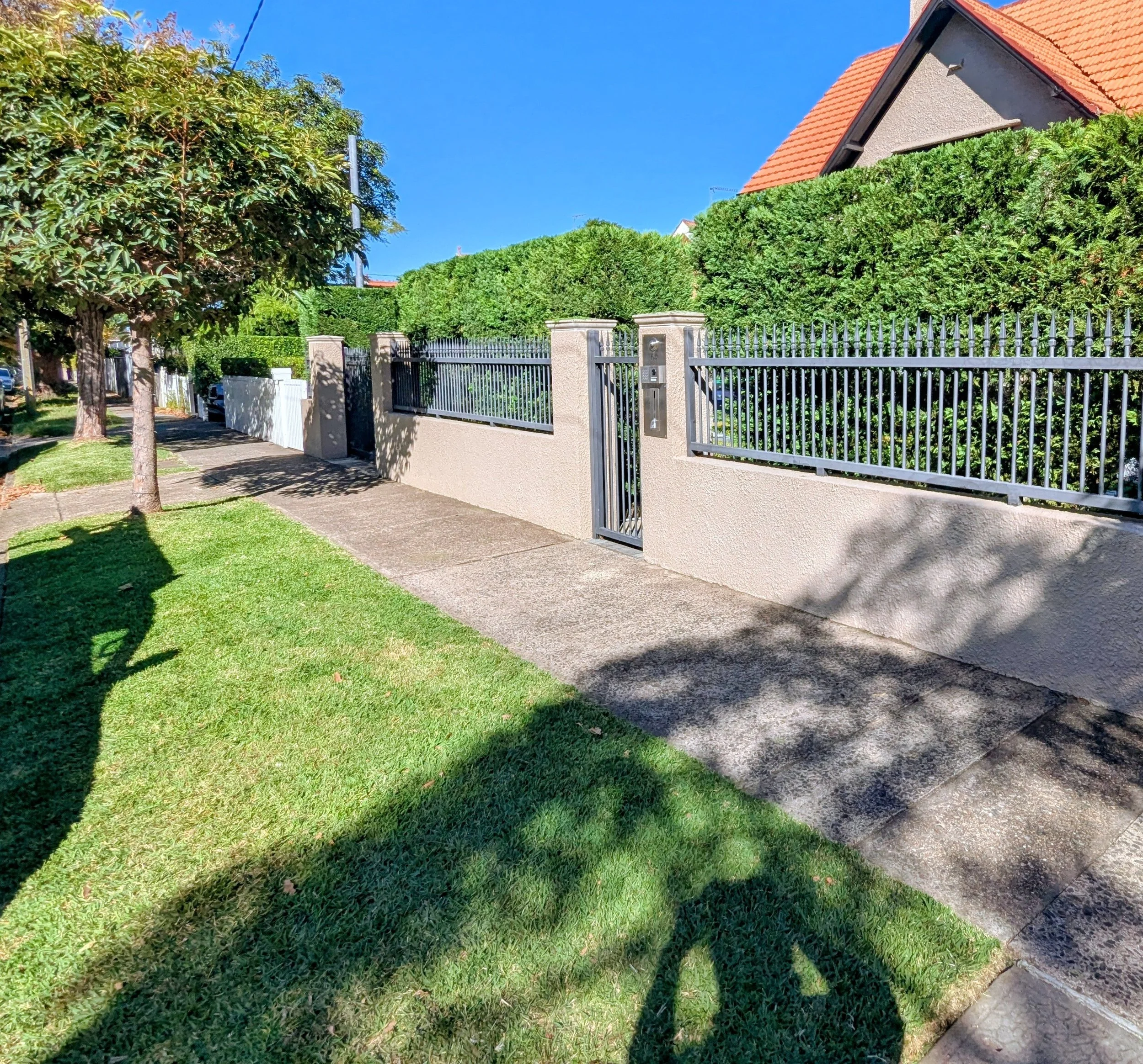Residential street scene with a sidewalk, green grass, and trees near a metal fence in front of a house with beige walls and a red-tiled roof, under a clear blue sky.