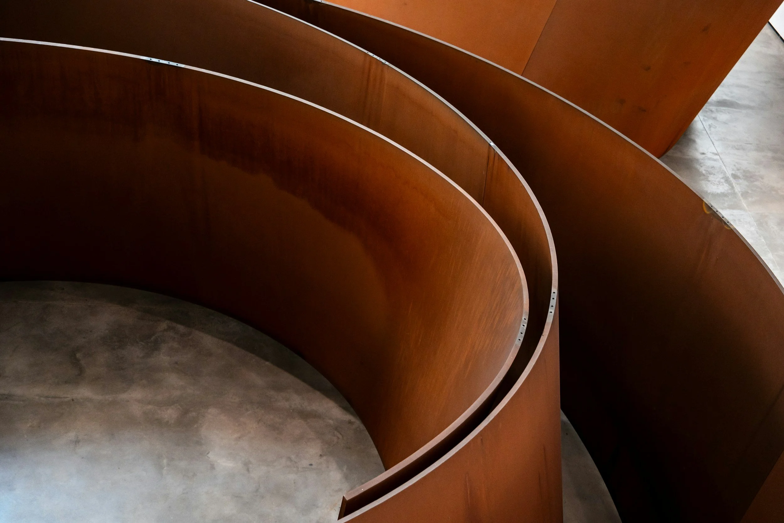 Close-up view of curved wooden panels with metallic edges, arranged in a spiral pattern on a concrete floor.