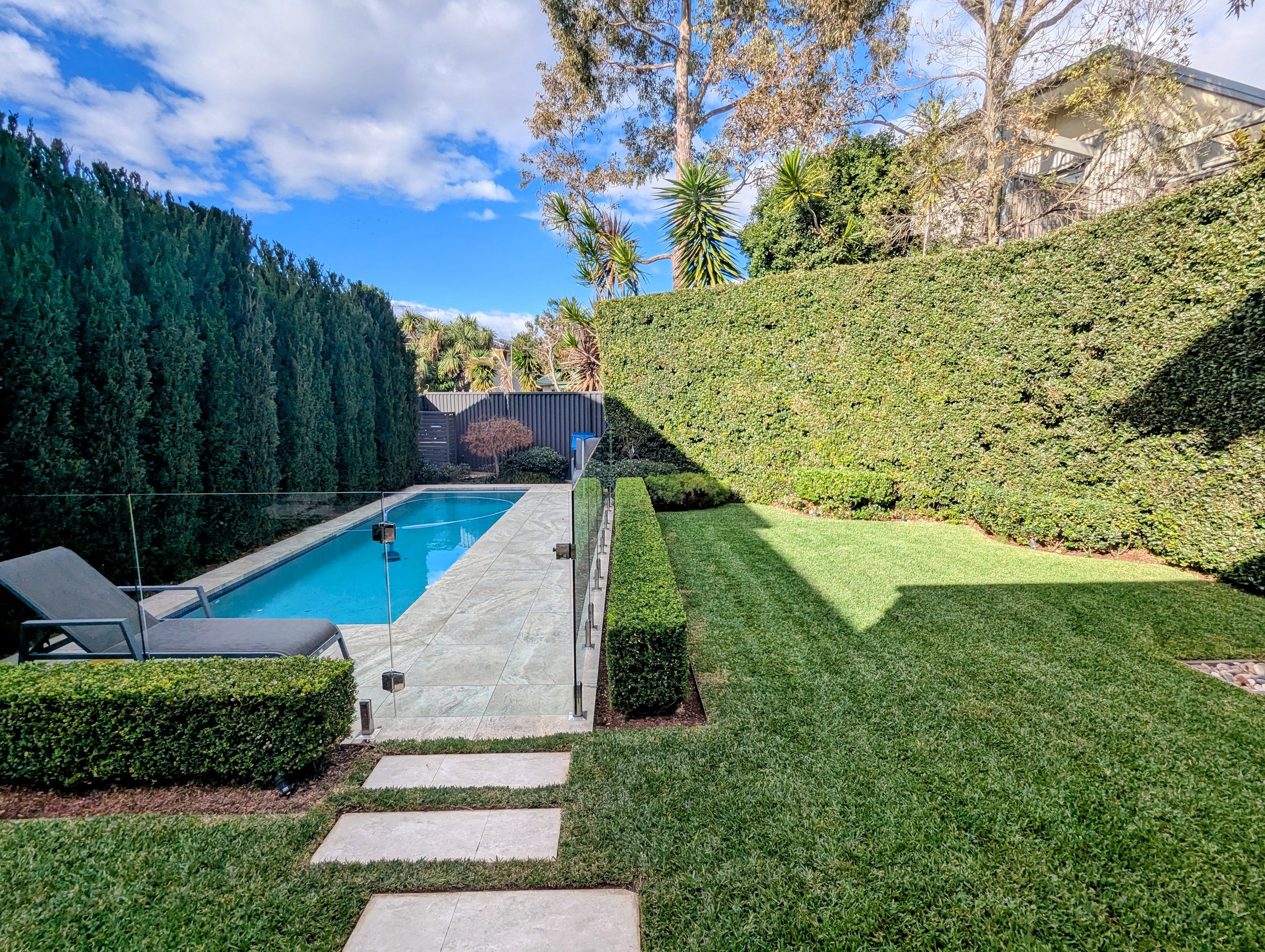 A backyard with a swimming pool, surrounded by a glass fence, green hedges, a lawn, and various trees, with a house in the background, under a partly cloudy sky.