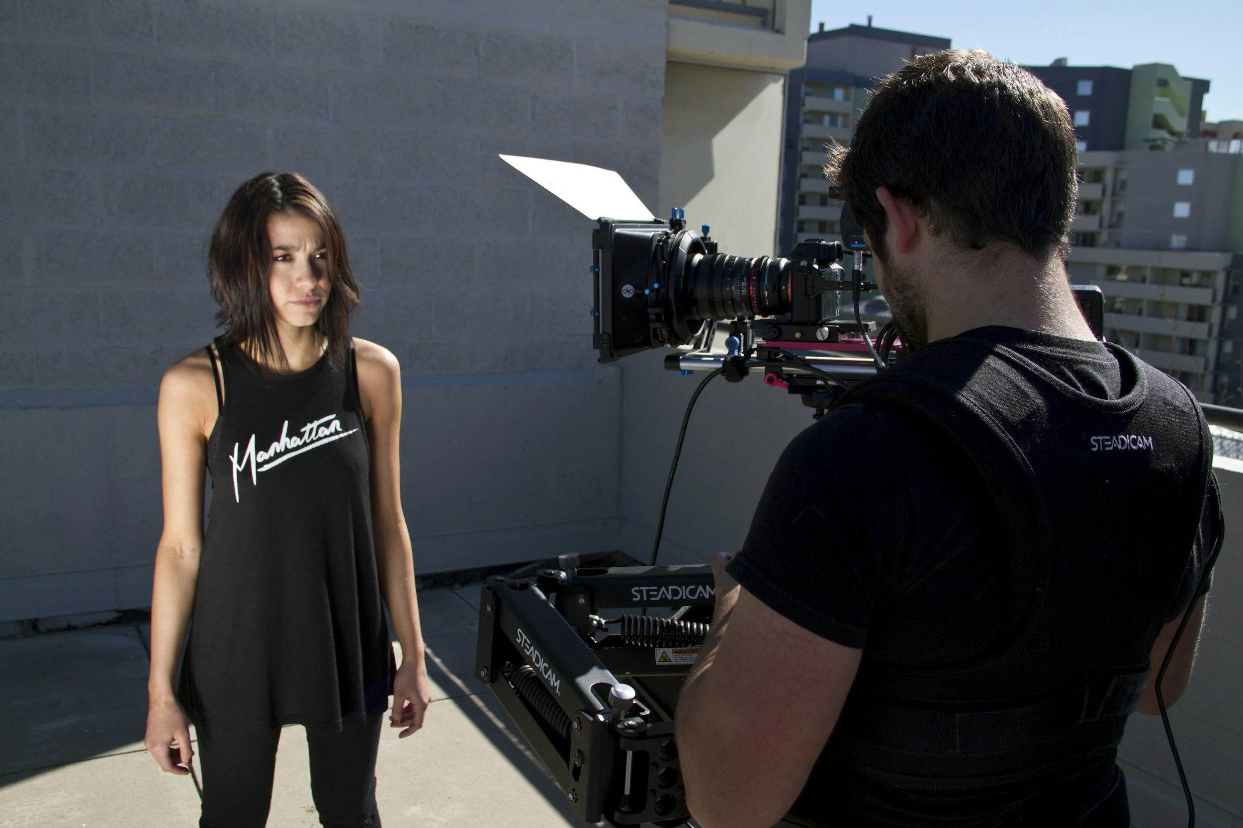 A woman with shoulder-length brown hair wearing a black tank top with 'Manhattan' written on it stands outdoors during the daytime. A man with dark hair and a beard, dressed in a black shirt, is operating a professional film camera on a steadycam rig, filming her. The background shows a cityscape with modern buildings and a clear blue sky.