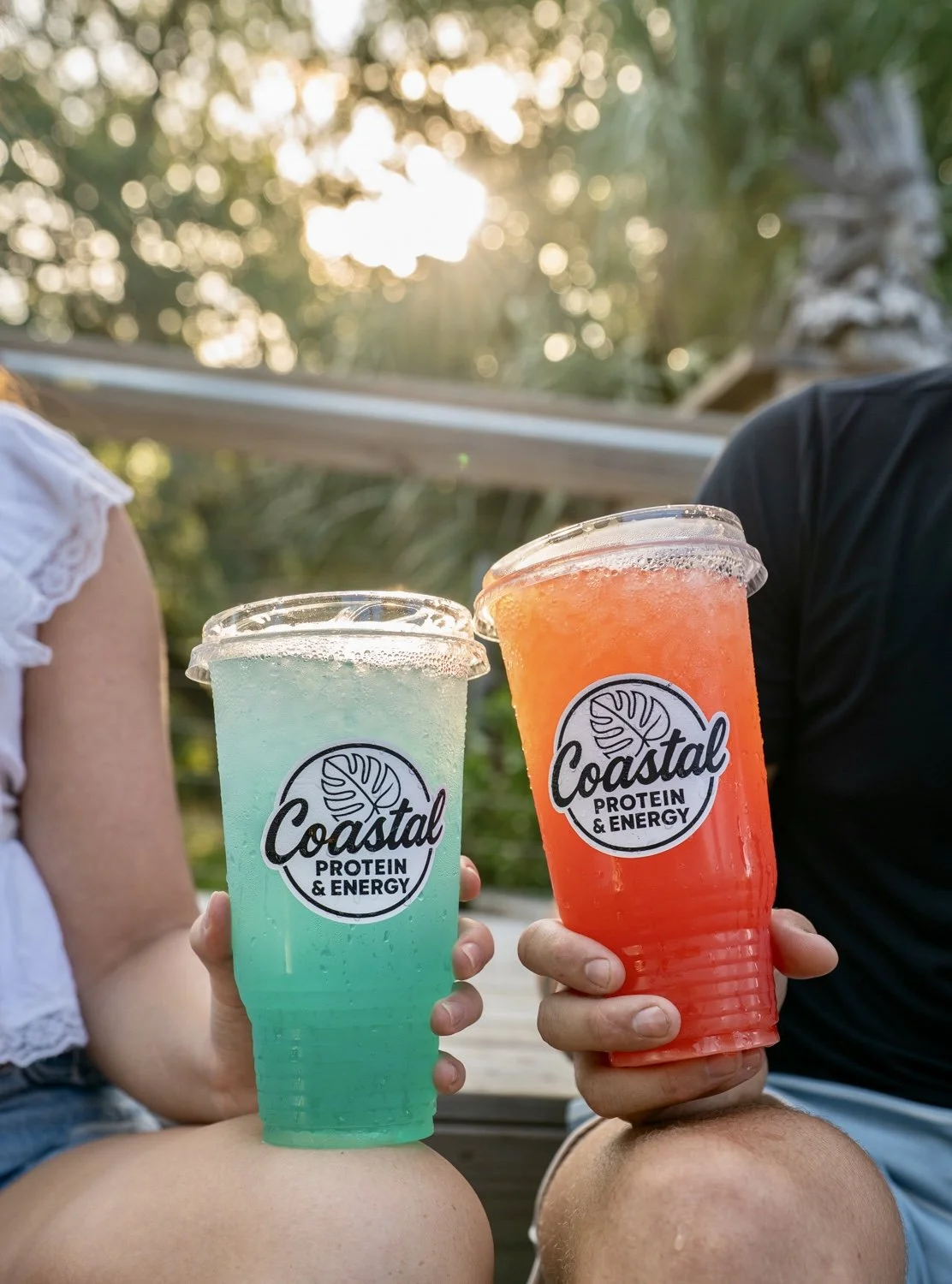 Two people holding Loaded Energy Teas in plastic cups with Coastal Protein & Energy logo, sitting outdoors in a park with sunlight filtering through trees.