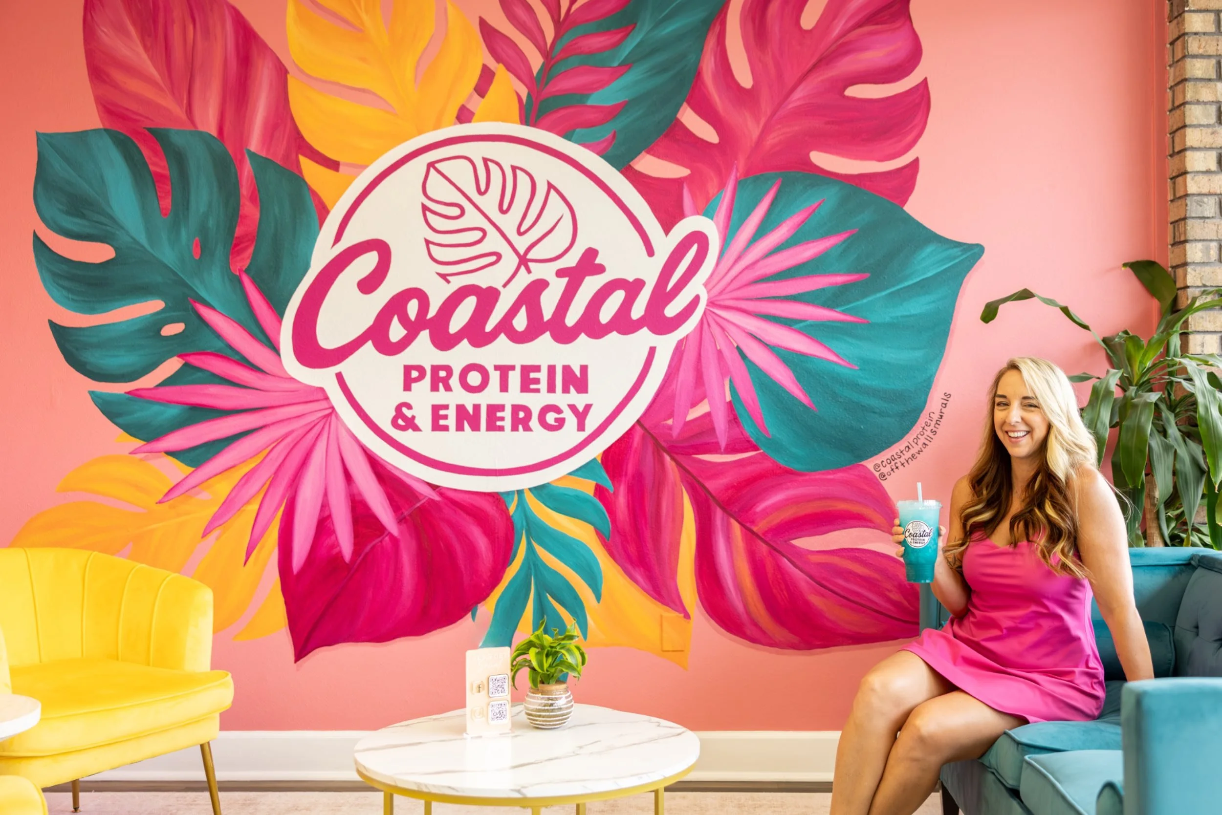 A woman in a pink dress holding a Loaded Tea with the Coastal Protein & Energy logo, sitting in a colorful cafe with a pink wall and tropical leaf mural.