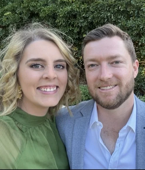 A smiling woman with curly blonde hair and a woman with a man with short dark hair and a beard taking a selfie outdoors with green trees in the background.