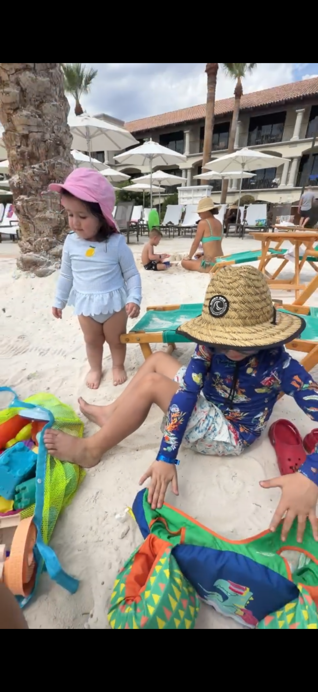 Children playing on a sandy beach with umbrellas and palm trees in the background. One girl in a pink hat and long-sleeved shirt stands near a seated boy with a large woven hat. Colorful beach bags and toys are scattered around.