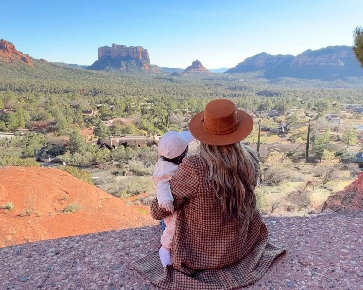 A woman with long wavy hair in a brown hat and coat kneels on a rocky ledge, holding a young child wearing a white hat and outfit, overlooking a scenic desert landscape with red rock formations and a small town.