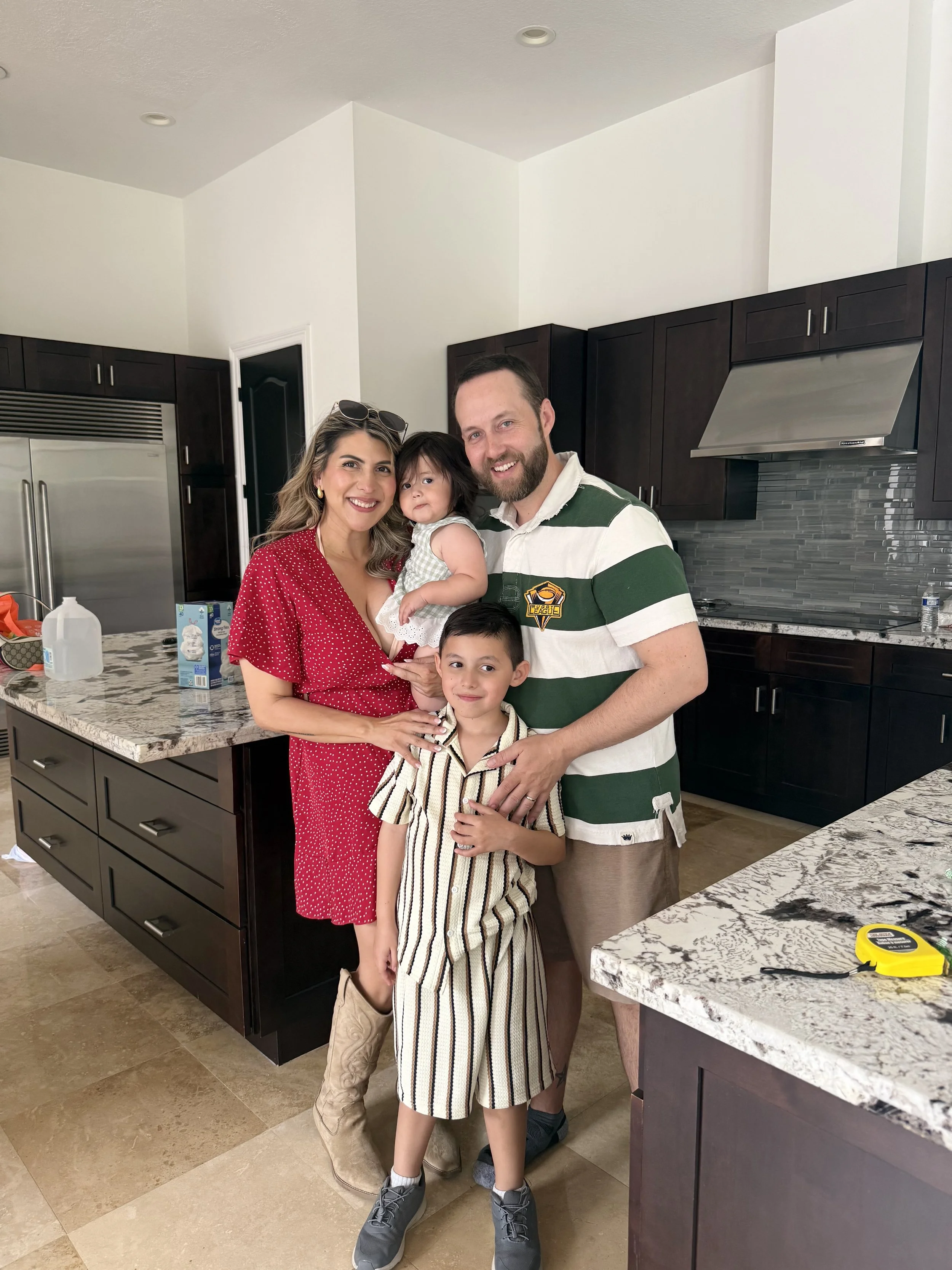 A family of four poses in a kitchen. The woman wears a red polka-dot dress, the man wears a green and white striped shirt, the girl in striped pajamas, and the boy in a plaid shirt and cowboy boots.