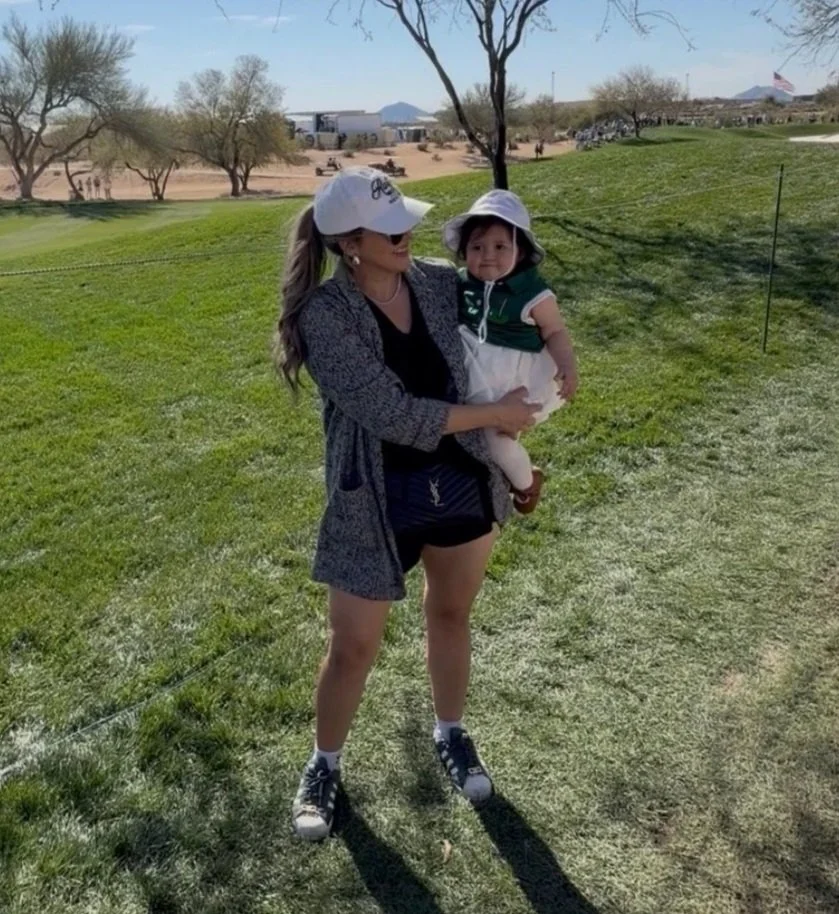 A woman holding a young girl outdoors in a park with green grass and trees, with mountains in the background.
