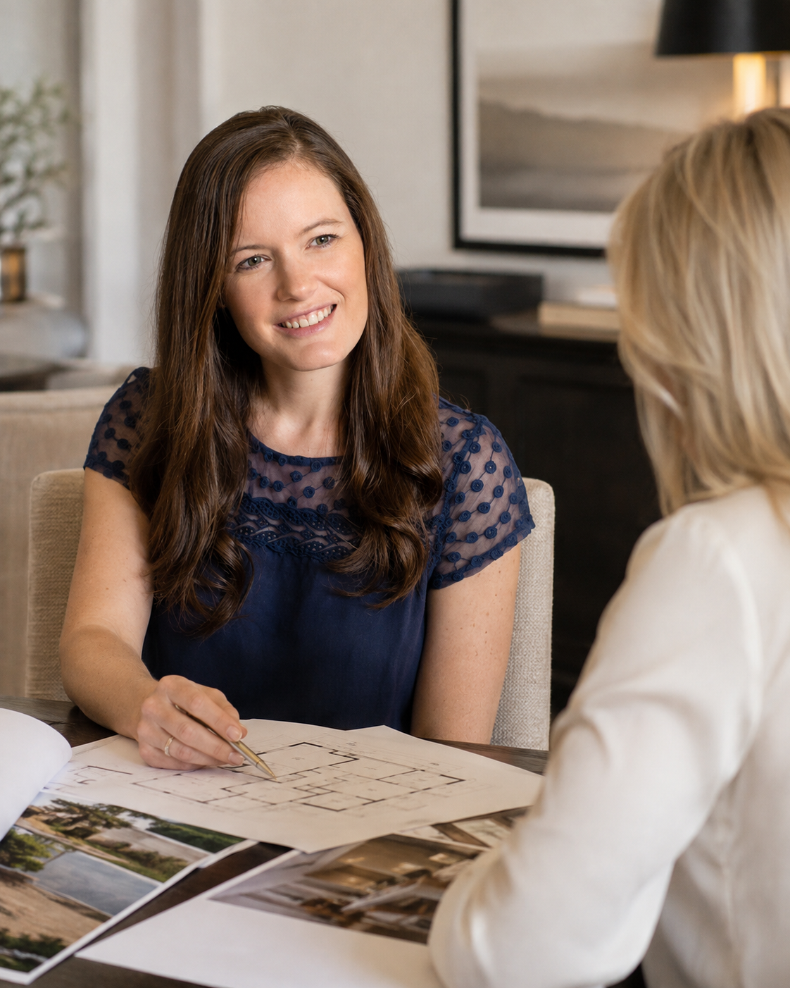 Two women sitting at a table having a discussion, with architectural plans and photographs spread out on the table.