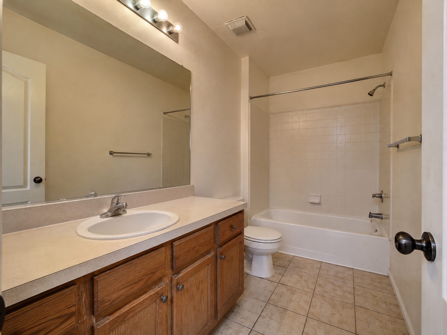 A bathroom with a large mirror above a wooden vanity with a sink, a toilet, and a bathtub with a shower. The wall tiles are white and the floor is tiled in beige.