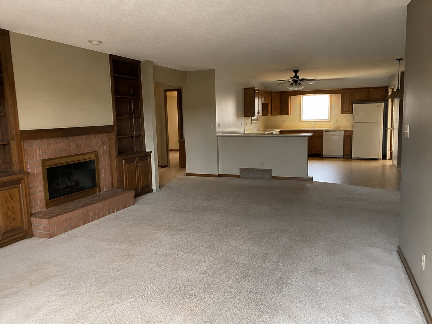 Empty living room with fireplace, built-in shelves, beige carpet, open kitchen with wooden cabinets, white appliances, and tiled floor.