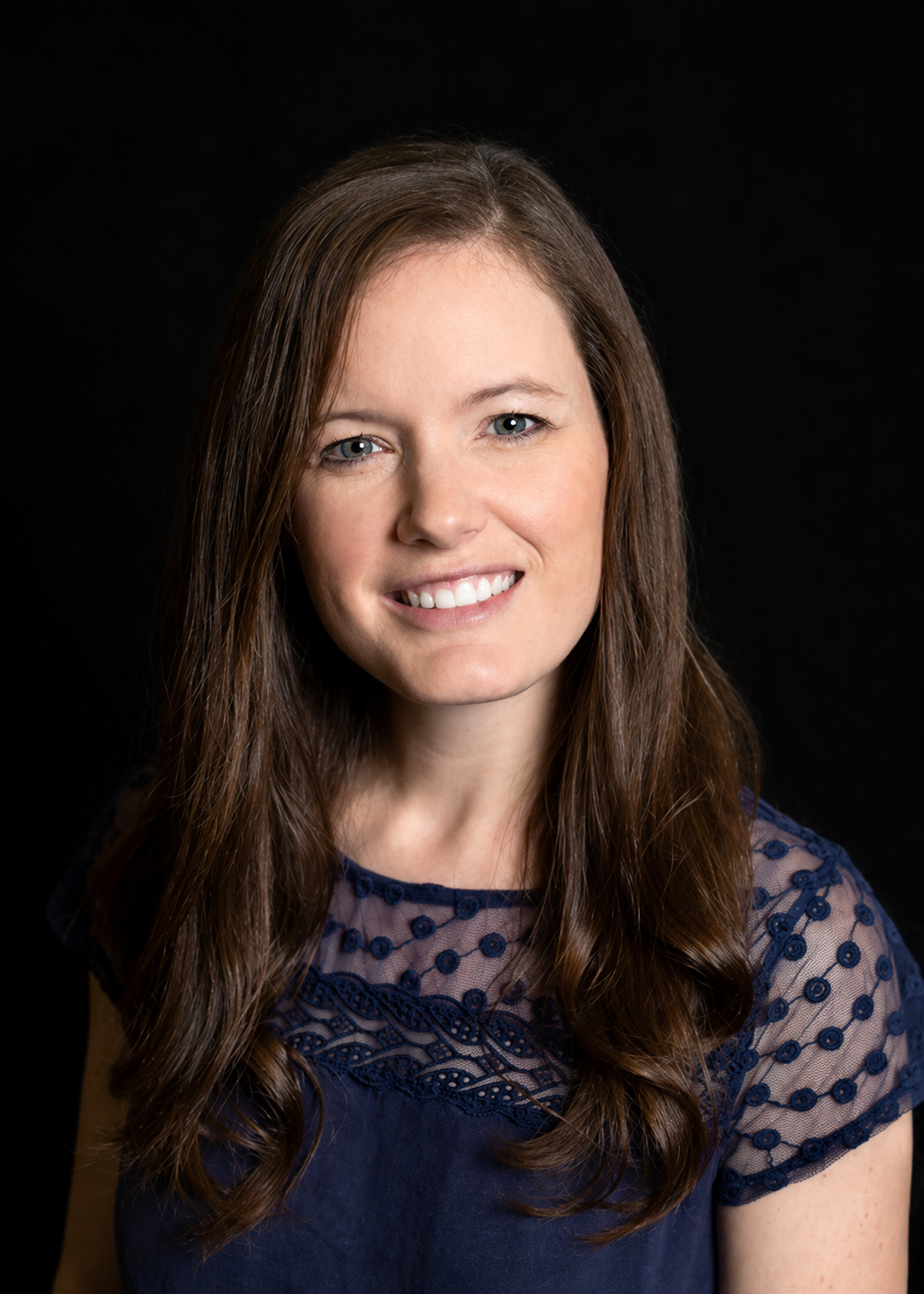 A woman with long brown hair, blue eyes, smiling, wearing a navy blue top with lace detail, against a black background.