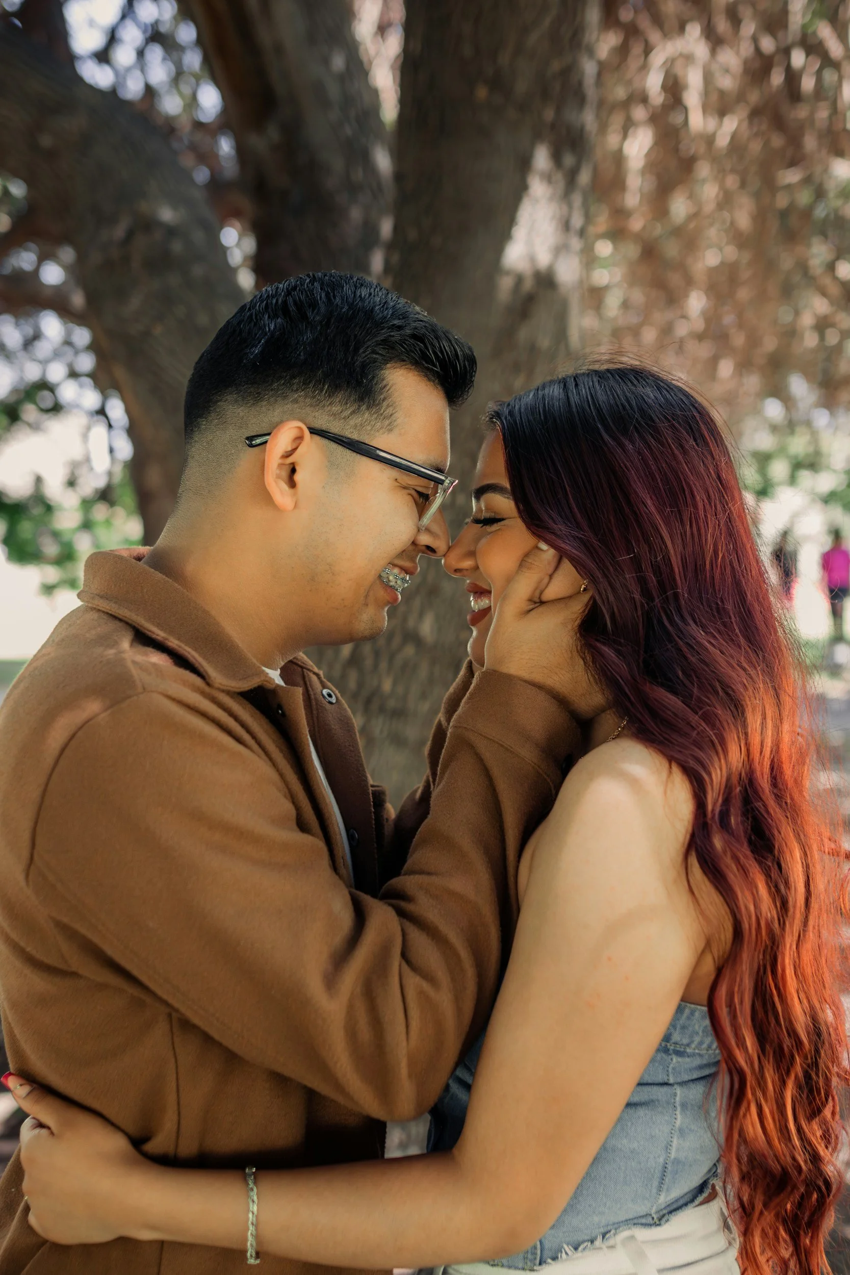 A young couple face to face, touching foreheads and smiling, outdoors near a large tree, with blurred background foliage.