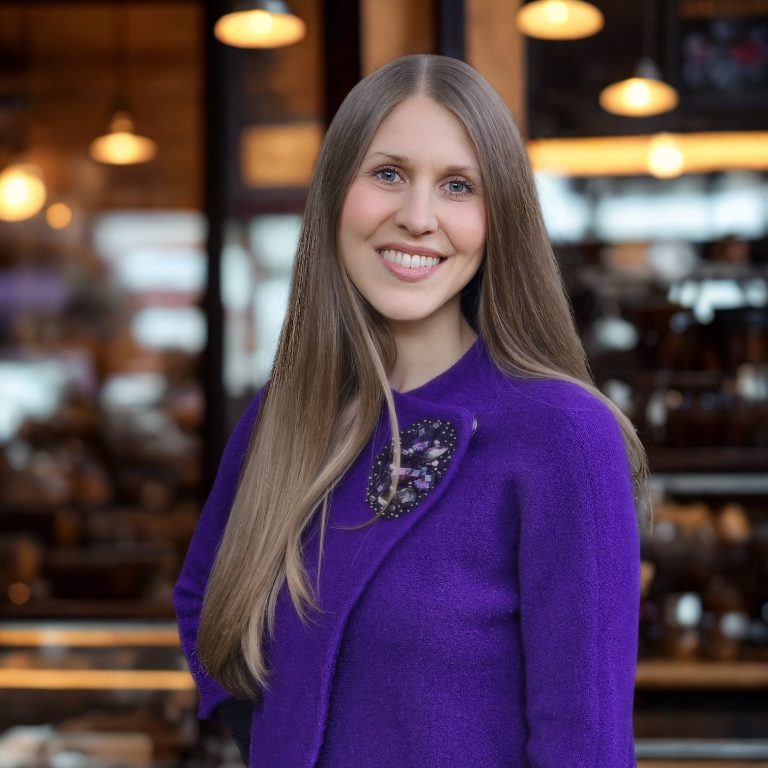 A smiling woman with long blonde hair wearing a purple sweater with a decorative patch on its shoulder, indoors with warm lighting and blurred background of a café or restaurant.