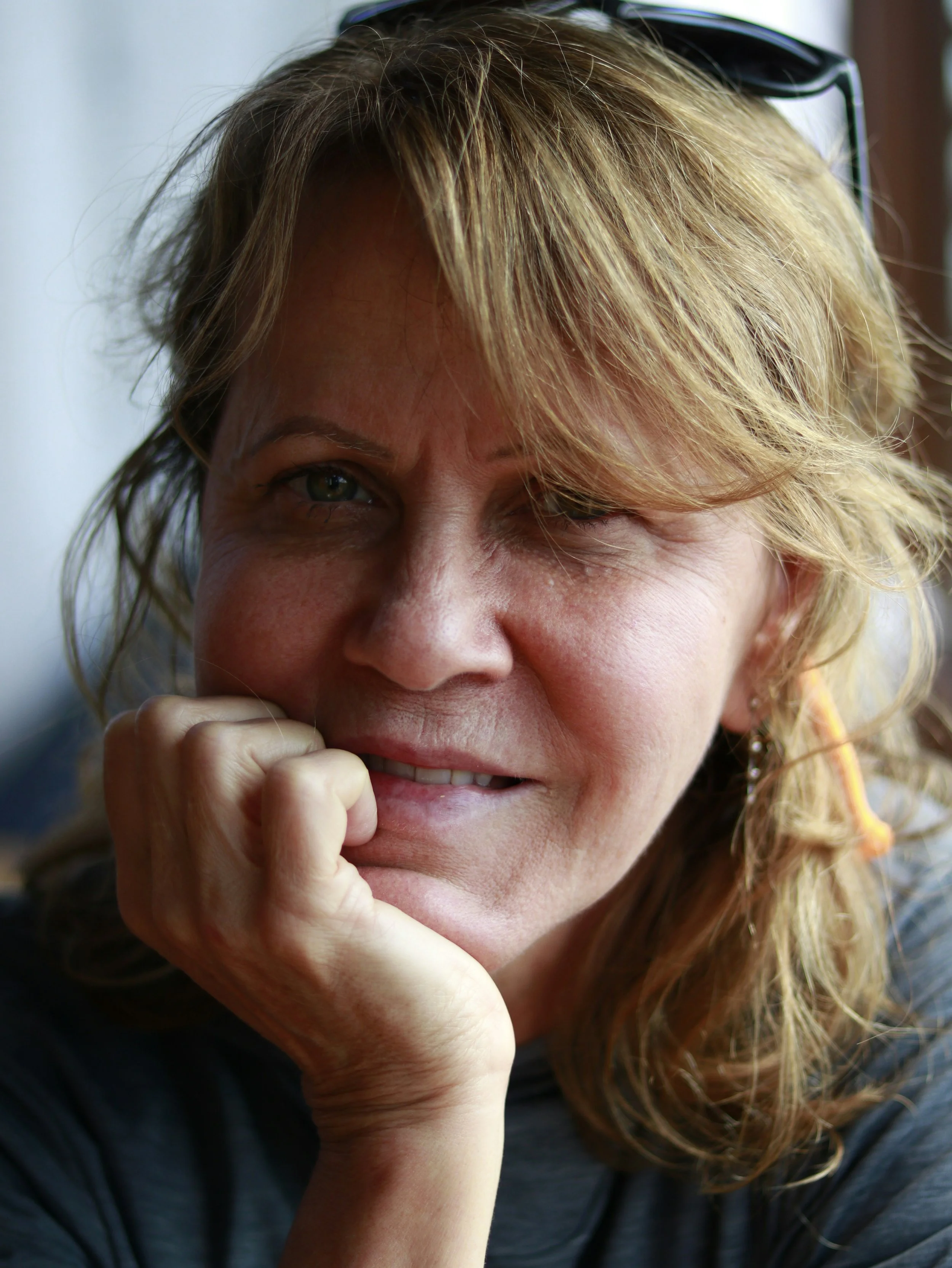 A woman with blonde hair and green eyes smiling and resting her face on her hand.
