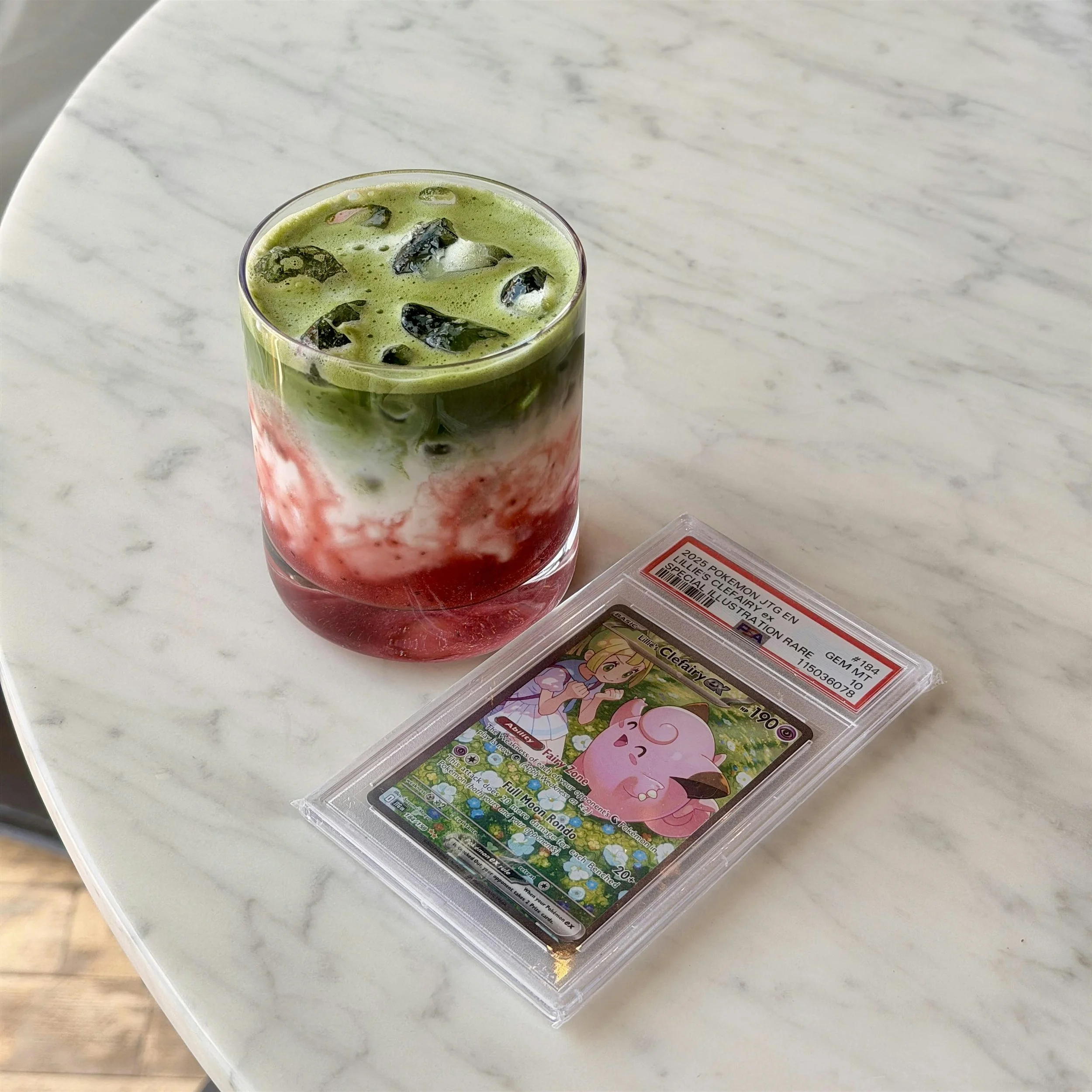 A Pokemon card inside a plastic case of a trainer kneeling next to Clefairy in a field of flowers on a marbled table with a glass cup of strawberry matcha latte next to it