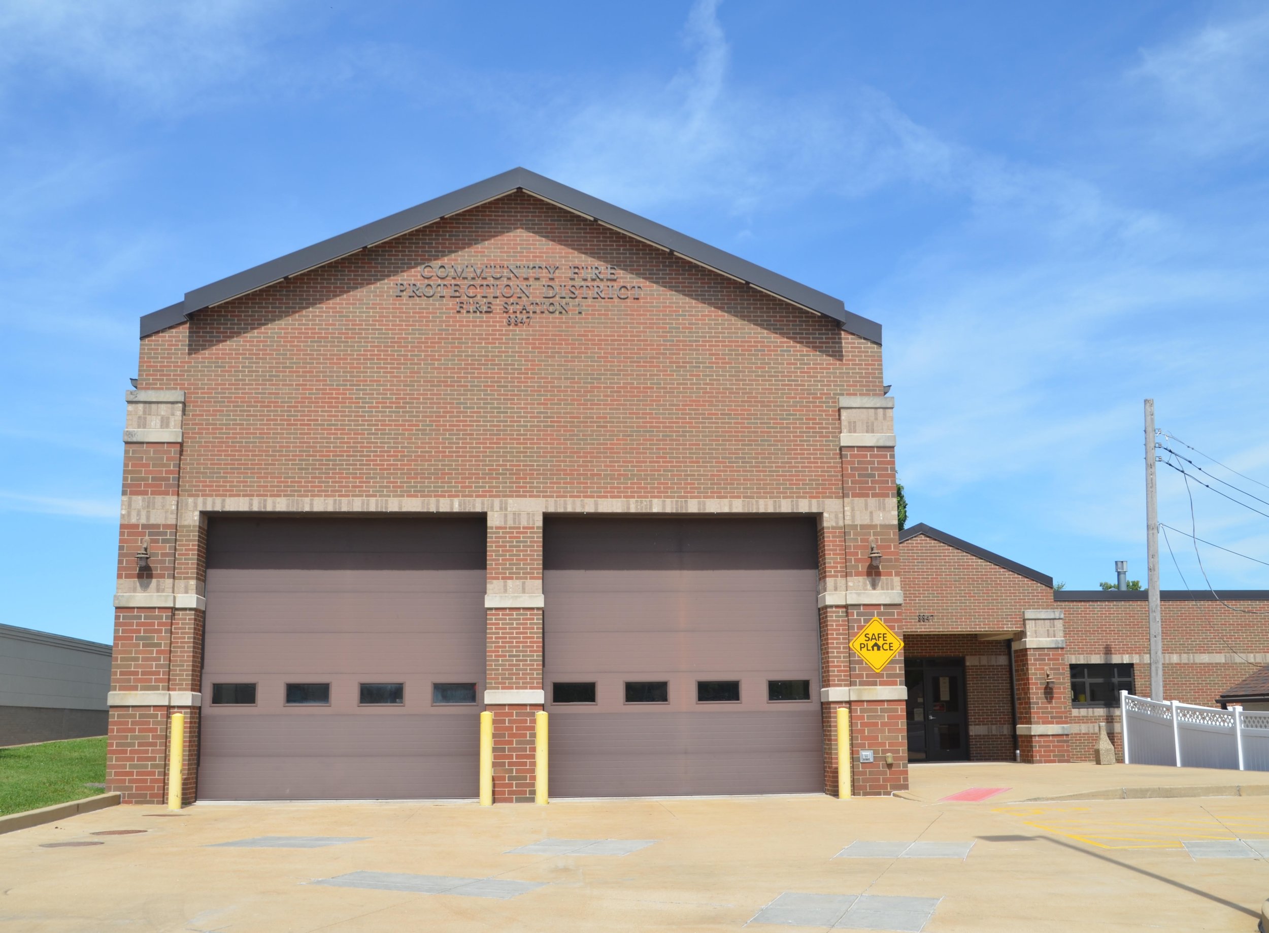 A brick fire station building with a sign that reads 'Community Fire Protection District Fire Station 1'. There are two garage doors in the front and a side entrance on the right. A yellow sign below the side entrance says 'Safe Place'.
