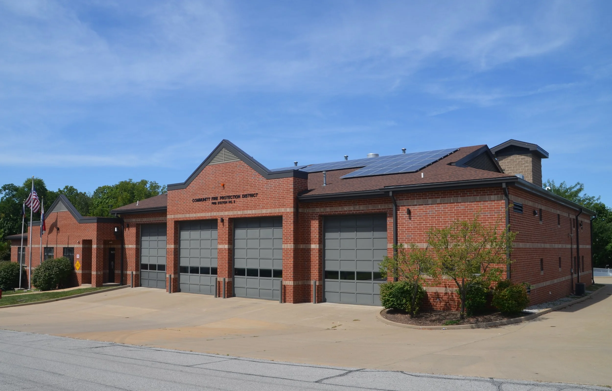 A red brick fire station with gray garage doors, solar panels on the roof, and a sign that reads 'Community Fire Protection District'. There are American flags and trees with green foliage in front of the building, under a blue sky with a few clouds.