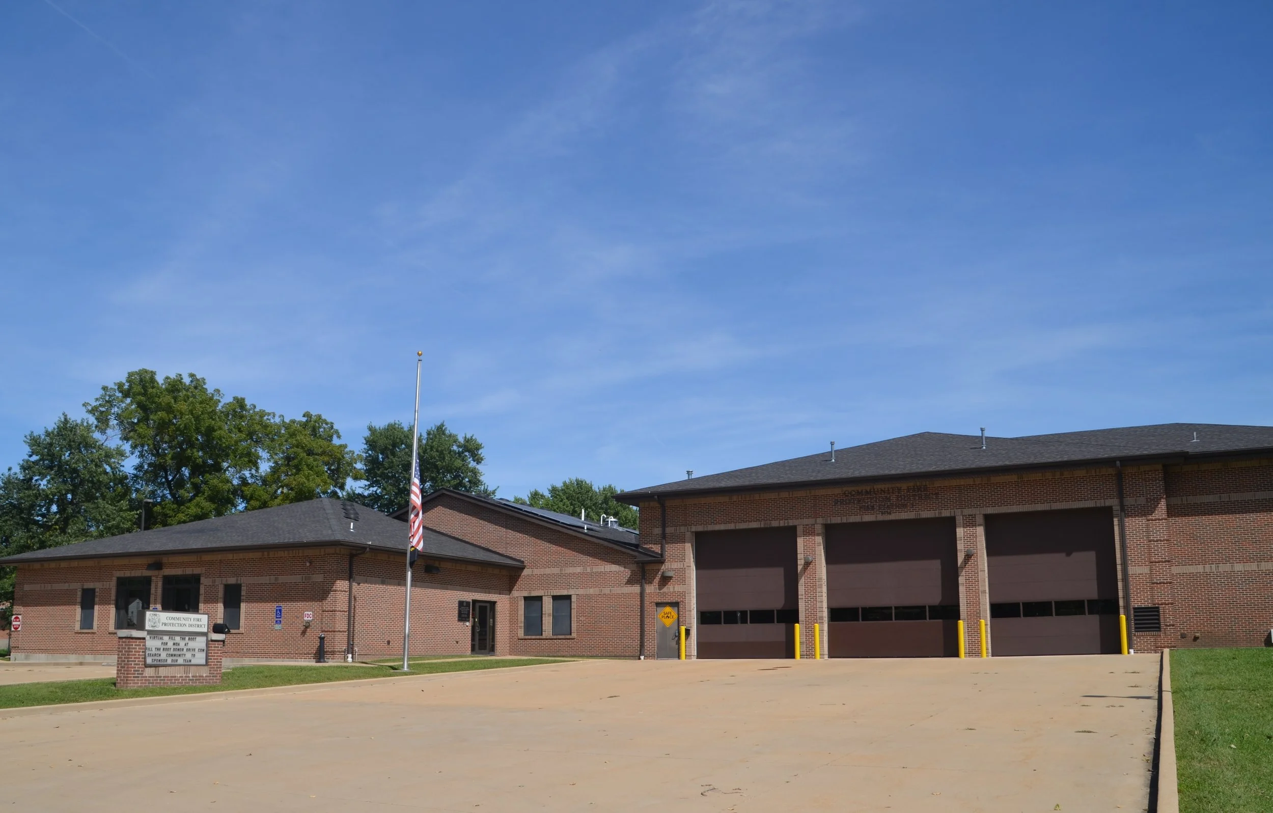 A brick fire station building with a flat roof and garage doors, American flag on a pole in front, trees in the background, and a clear blue sky overhead.