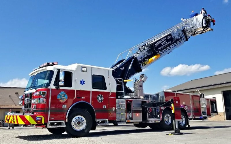 A community fire truck with an extended ladder parked outdoors on pavement, with a building and blue sky in the background.