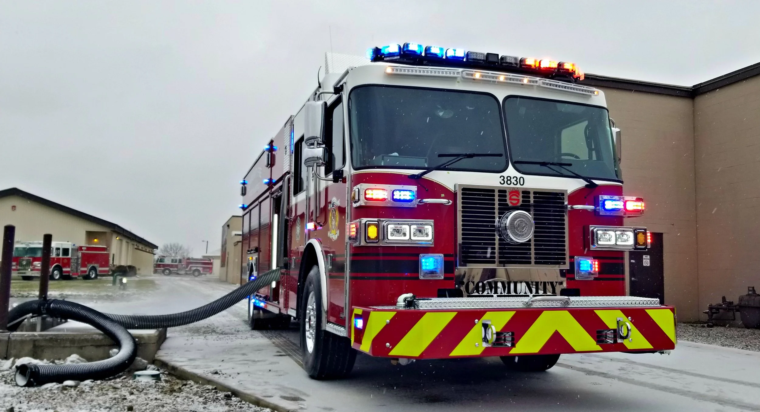 A red fire truck with blue and red emergency lights parked outdoors on a snowy day, with several other fire trucks and a beige building in the background.