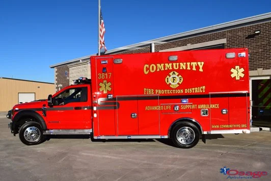 Red community ambulance truck parked in front of a building with an American flag in the background.
