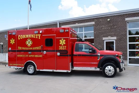 Red community fire protection district ambulance vehicle parked outside a brick building with large windows and a fire station door.