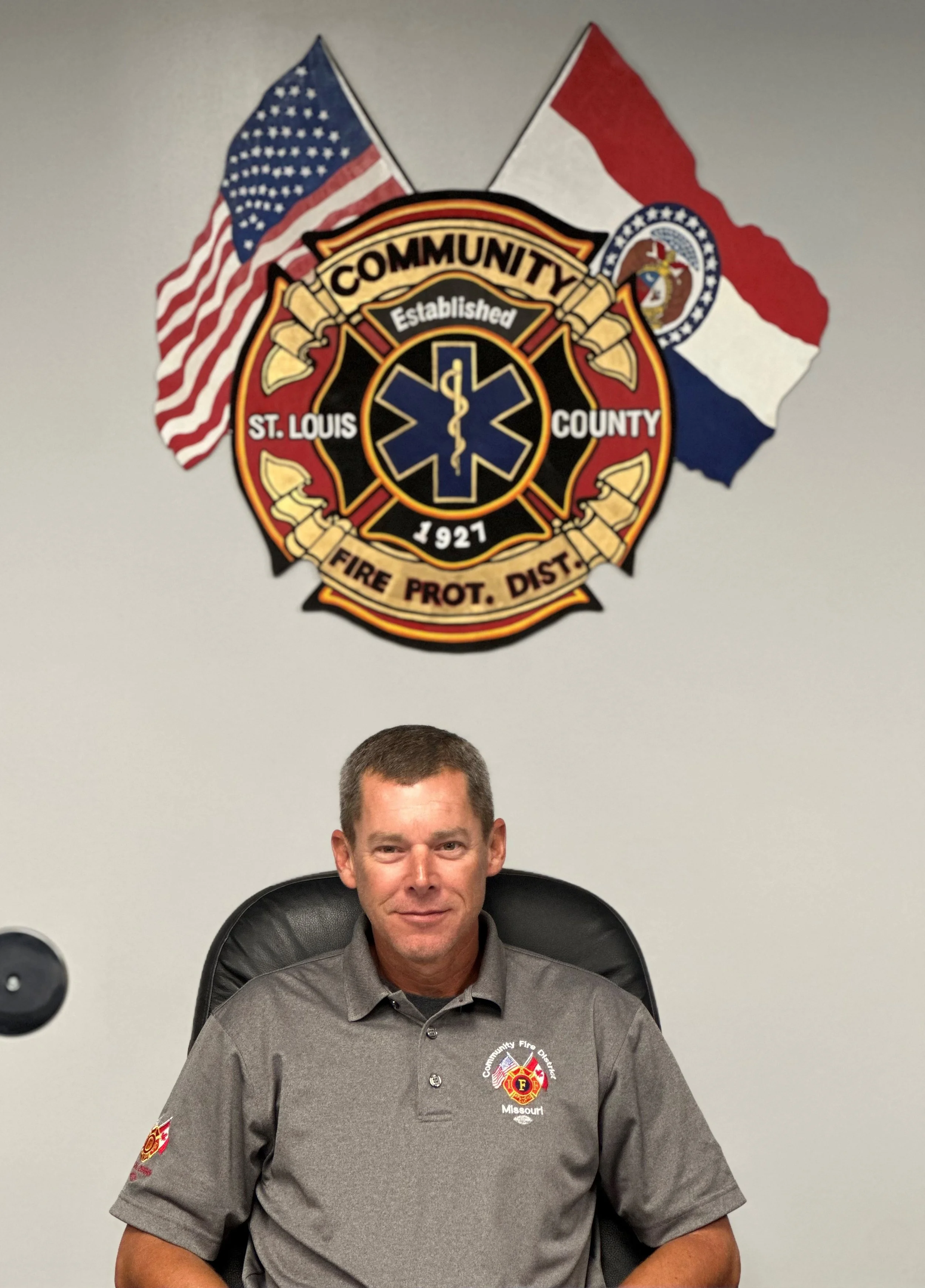 A man sitting in a black leather office chair in front of a wall with a fire department emblem and two American flags. The emblem reads 'Community Established 1921 St. Louis County Fire Prot. Dist.' The man is wearing a gray firefighter uniform shirt with patches, and has short dark hair, smiling slightly.