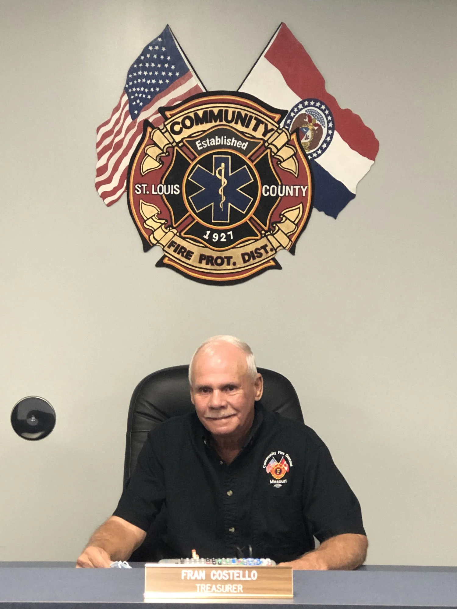 A man sitting at a table with a nameplate that says 'FRAN COSTELLO TREASURER.' He is wearing a black shirt with a fire department badge and the word 'Missouri' on it. Behind him on the wall are two flags, one American and one Missouri state flag, and a firefighter emblem with the words 'Community' and 'St. Louis County' along with the year 1927.