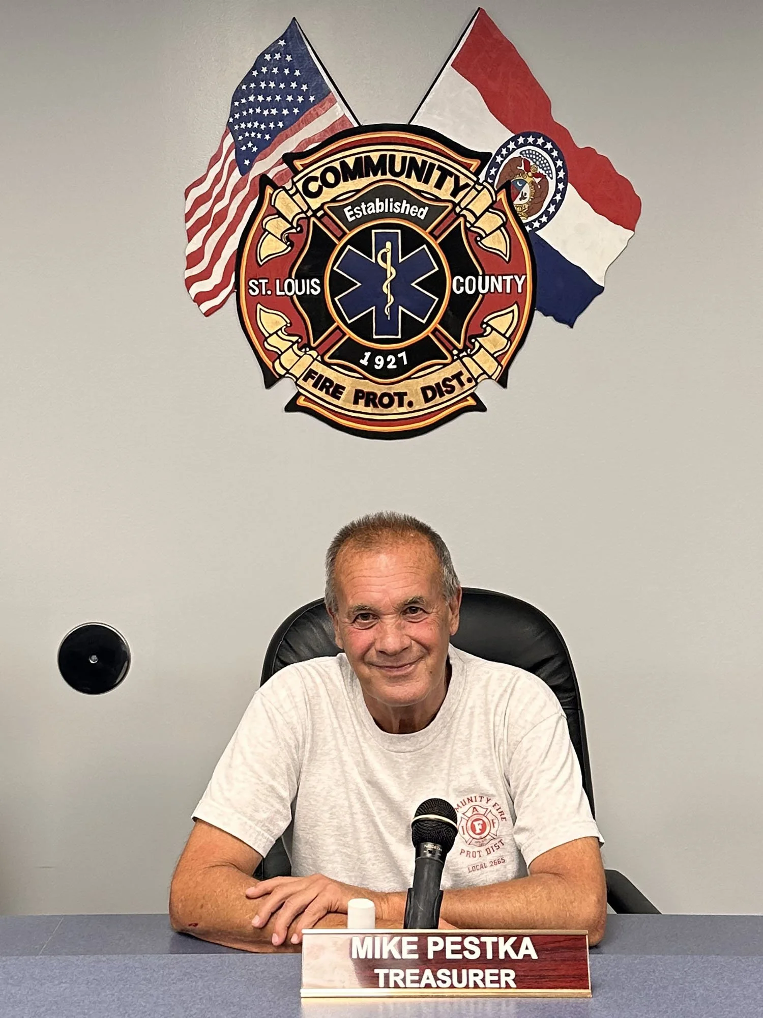 A man with gray hair sitting at a desk in front of a microphone. He is wearing a light-colored T-shirt and smiling. Behind him, there is a shield emblem for St. Louis County Fire Department. The nameplate on the desk reads 'Mike Pestka, Treasurer '.
